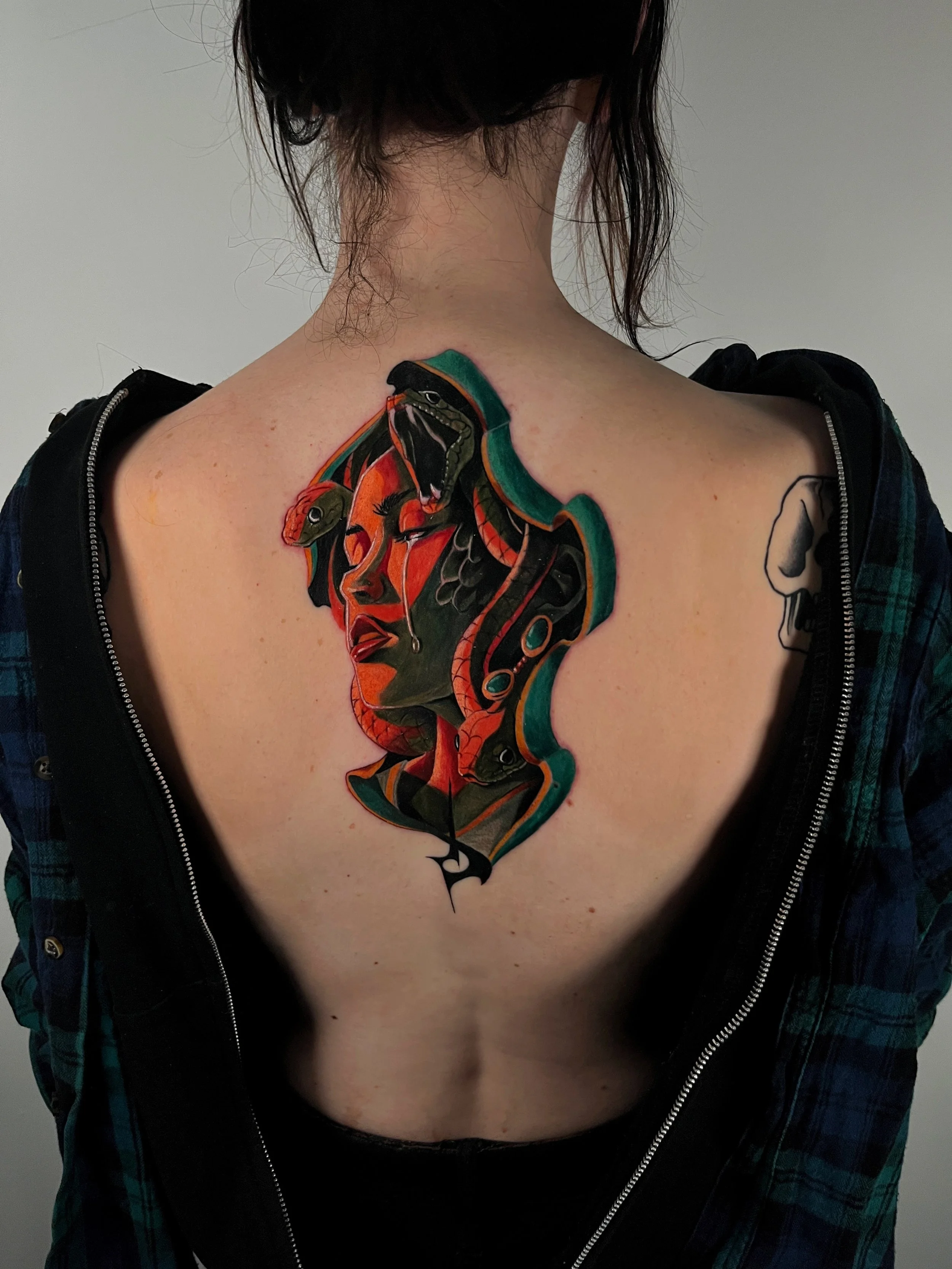 A tattoo on the upper back depicting a snake's head and a woman in profile in a traditional style, with bright colors.
