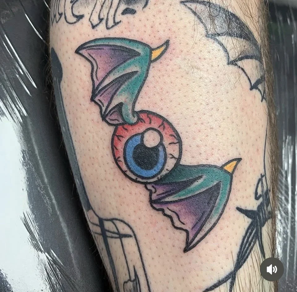 A tattoo of an eye with bat wings in bright colors on the skin.