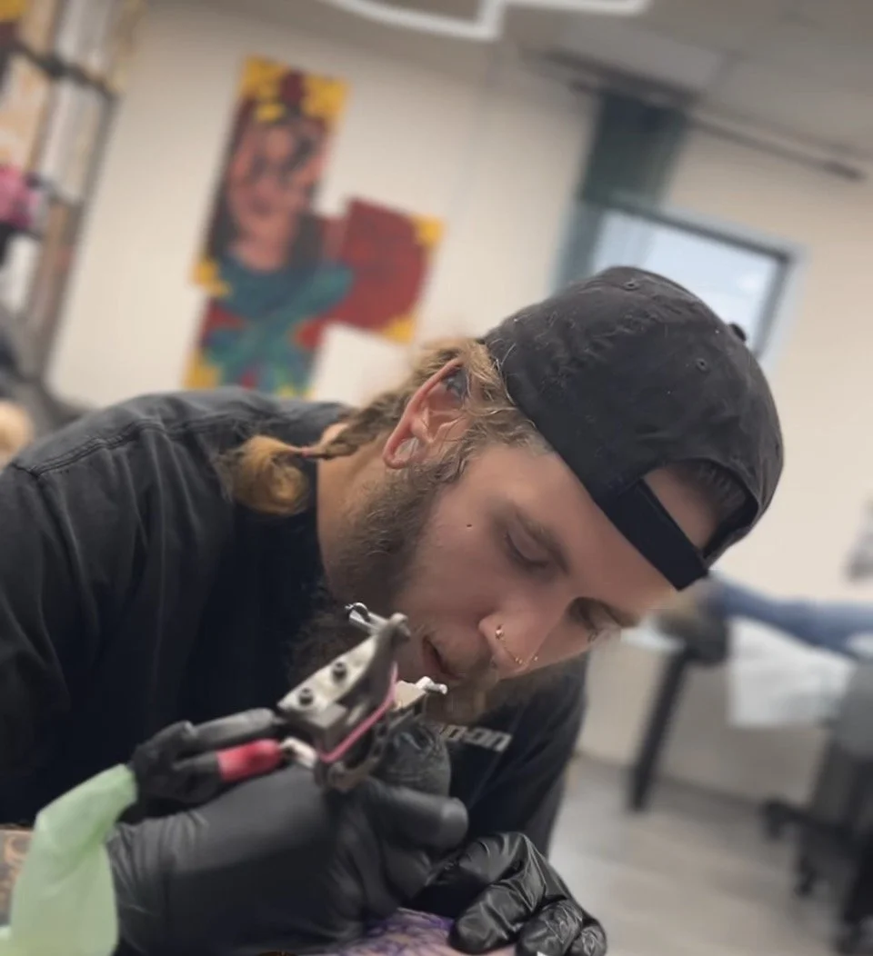 A man wearing a black cap, with piercings and a tattoo, using an electric tool in a workshop, with a colorful painting in the background.