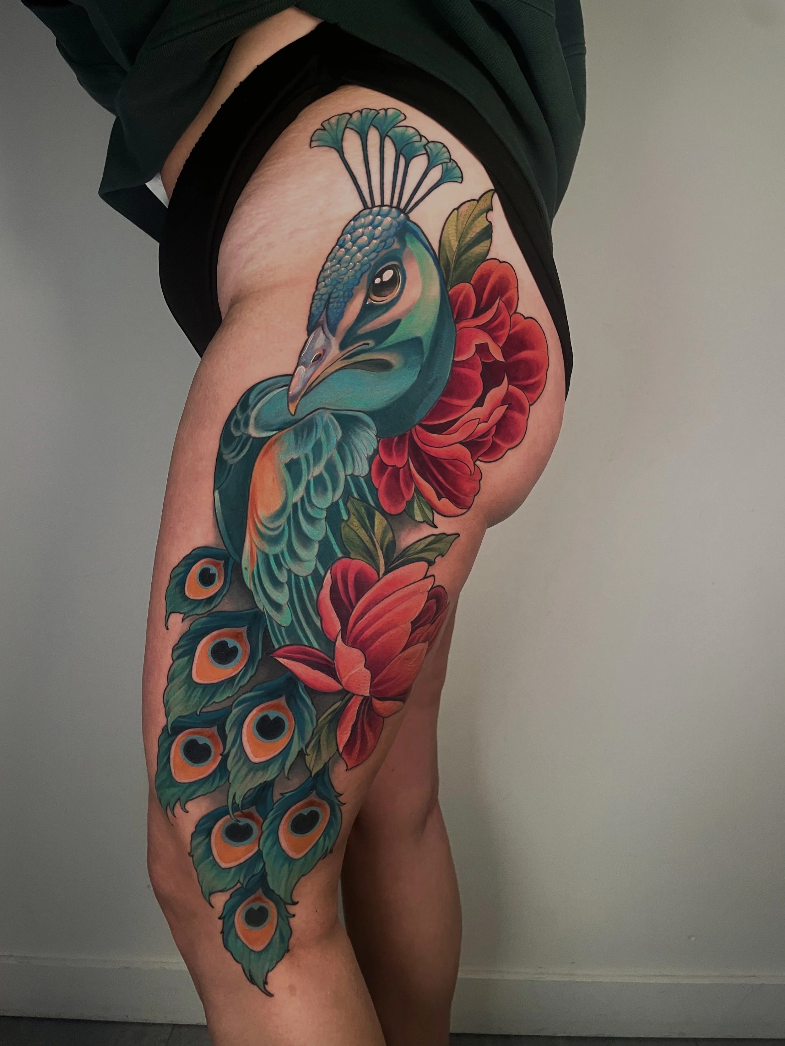 Tatouage coloré d'un paon et de fleurs rouges sur la cuisse d'une personne.