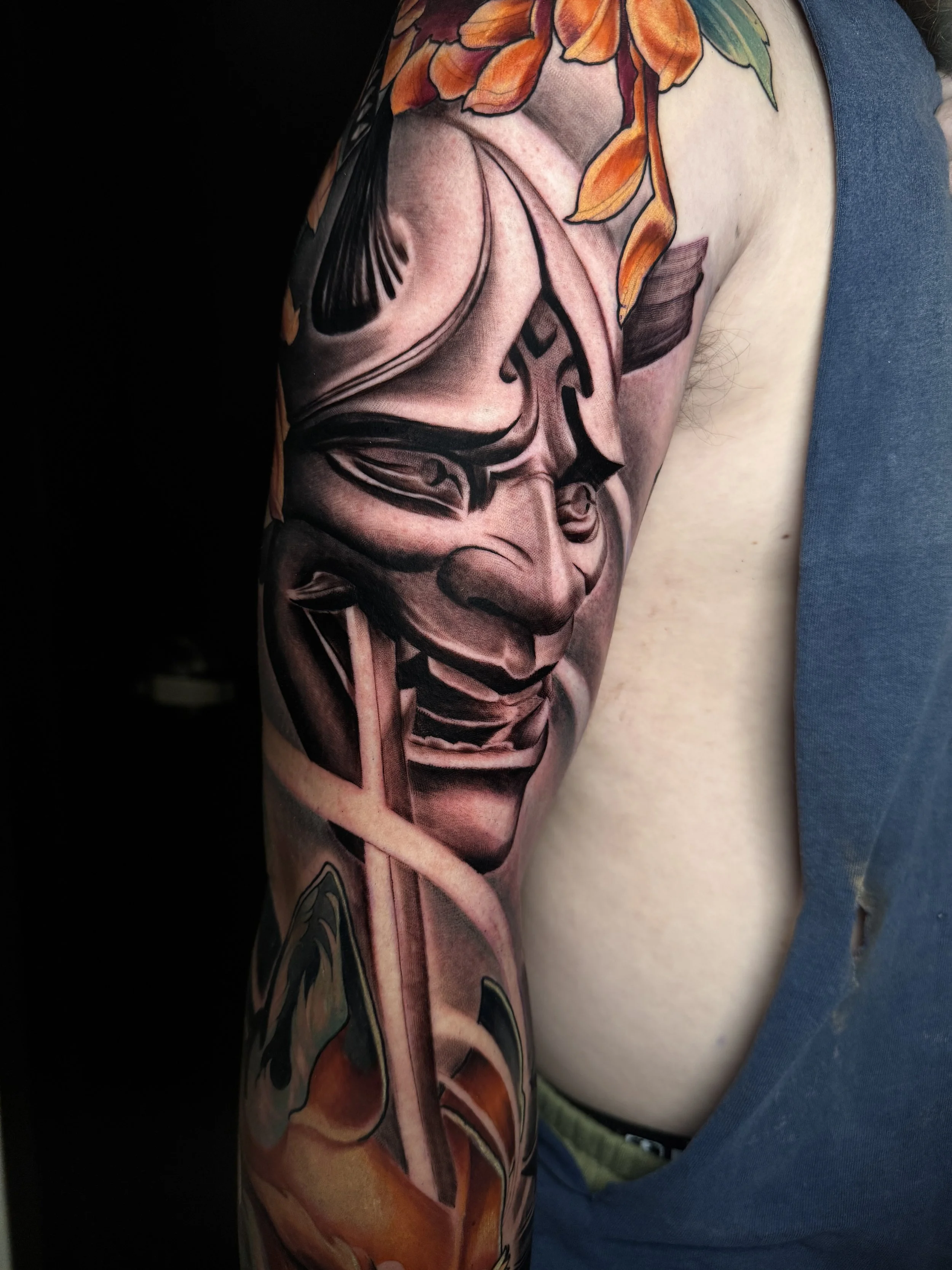 A detailed tattoo depicting a human face with floral and natural elements, located on the arm of a person wearing black clothing and a gray T-shirt.