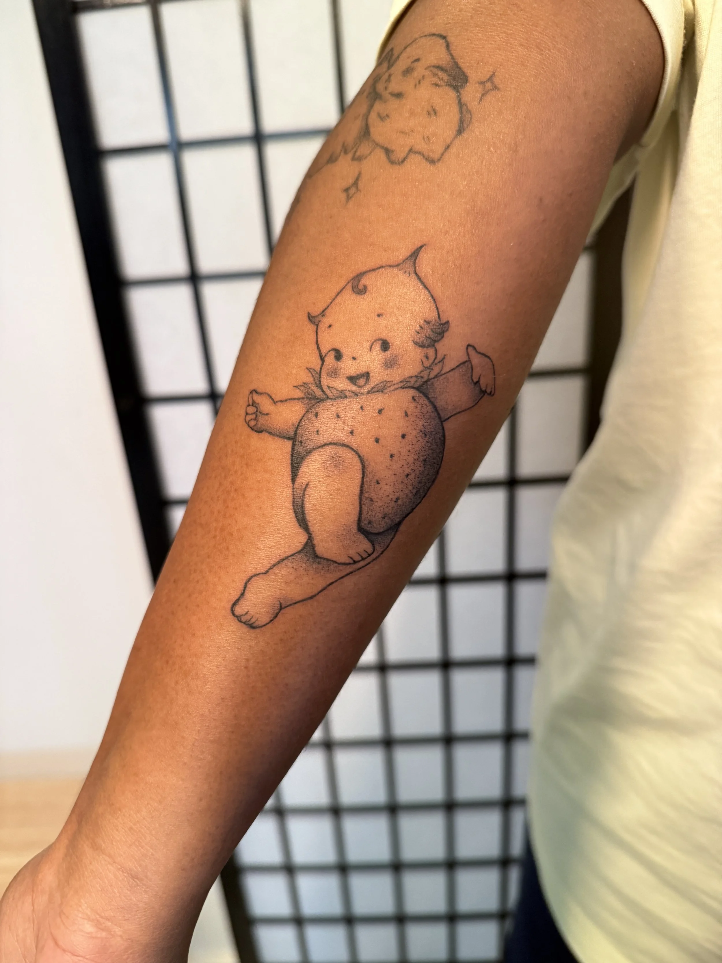 Tattoo of a cute character with pointed ears, a happy smile, and a round body with spots, sitting with one arm raised.