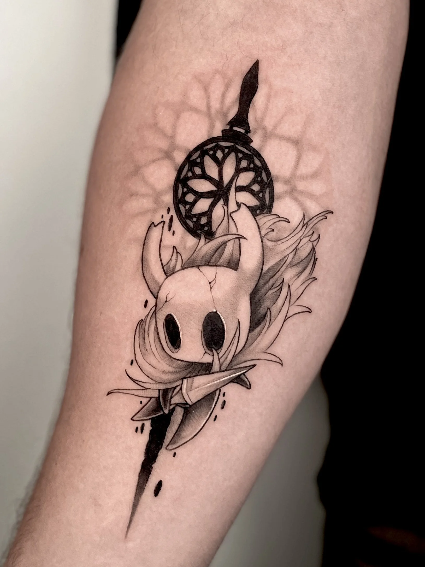 Black and white tattoo of a small animal with large eyes, a small snout, and large ears, surrounded by flowers and a spear with an upward-pointing tip, with an ornate circle in the background.