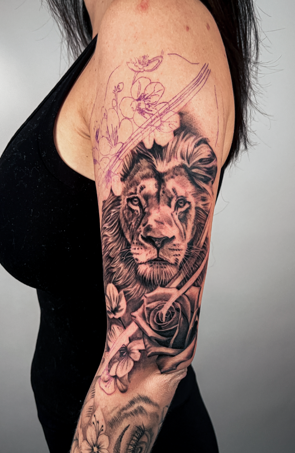 Tattoo of a realistic lion surrounded by flowers, including a rose and butterflies, on a woman's arm.