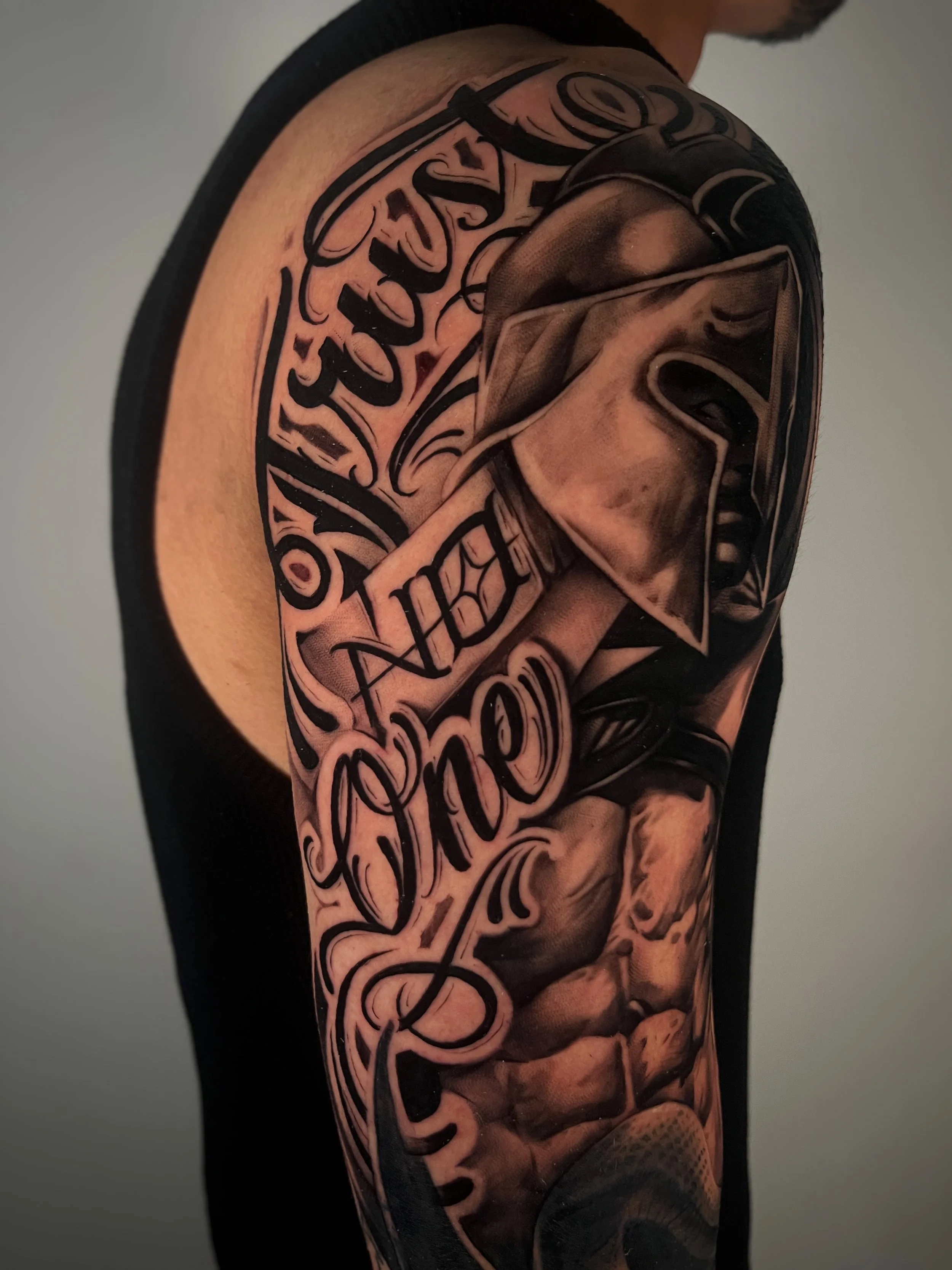 A tattoo of a Greek Atlas head in black and gray on a man's shoulder, with words in cursive script incorporated into the design.