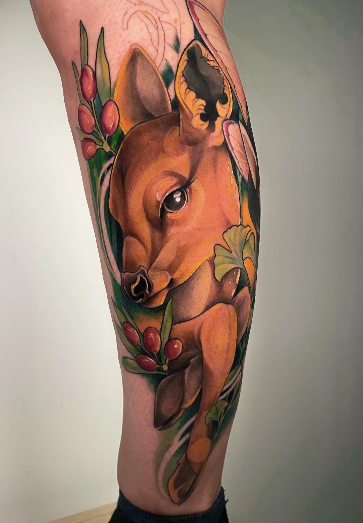 Tatouage réaliste d'un faon couché entouré de feuilles, de branches et de fleurs colorées sur le bras.