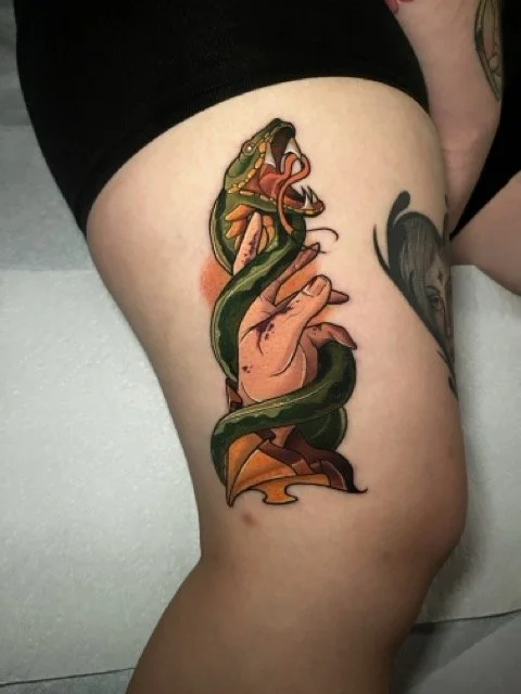Tattoo of a hand emerging from a snake, with a snake's head wrapped around it, on a person's thigh.