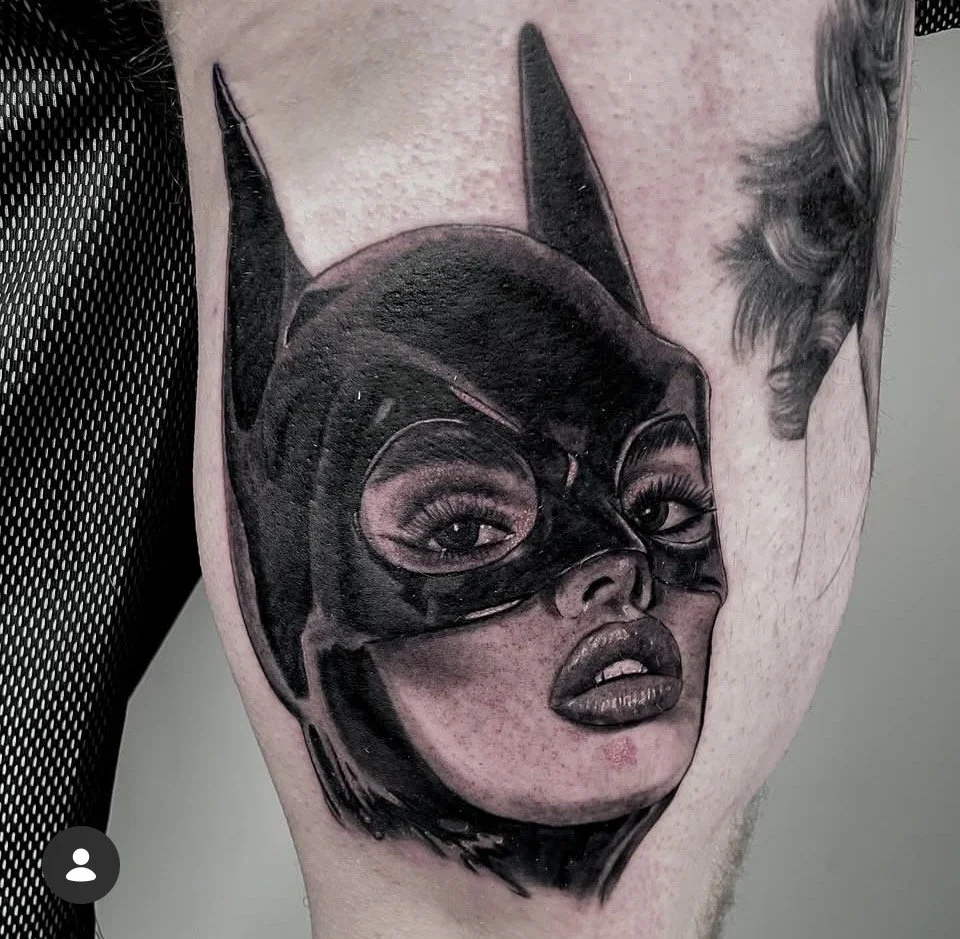 A black-and-gray tattoo depicting a woman's face wearing a Batman mask, with realistic details on both the face and the mask.