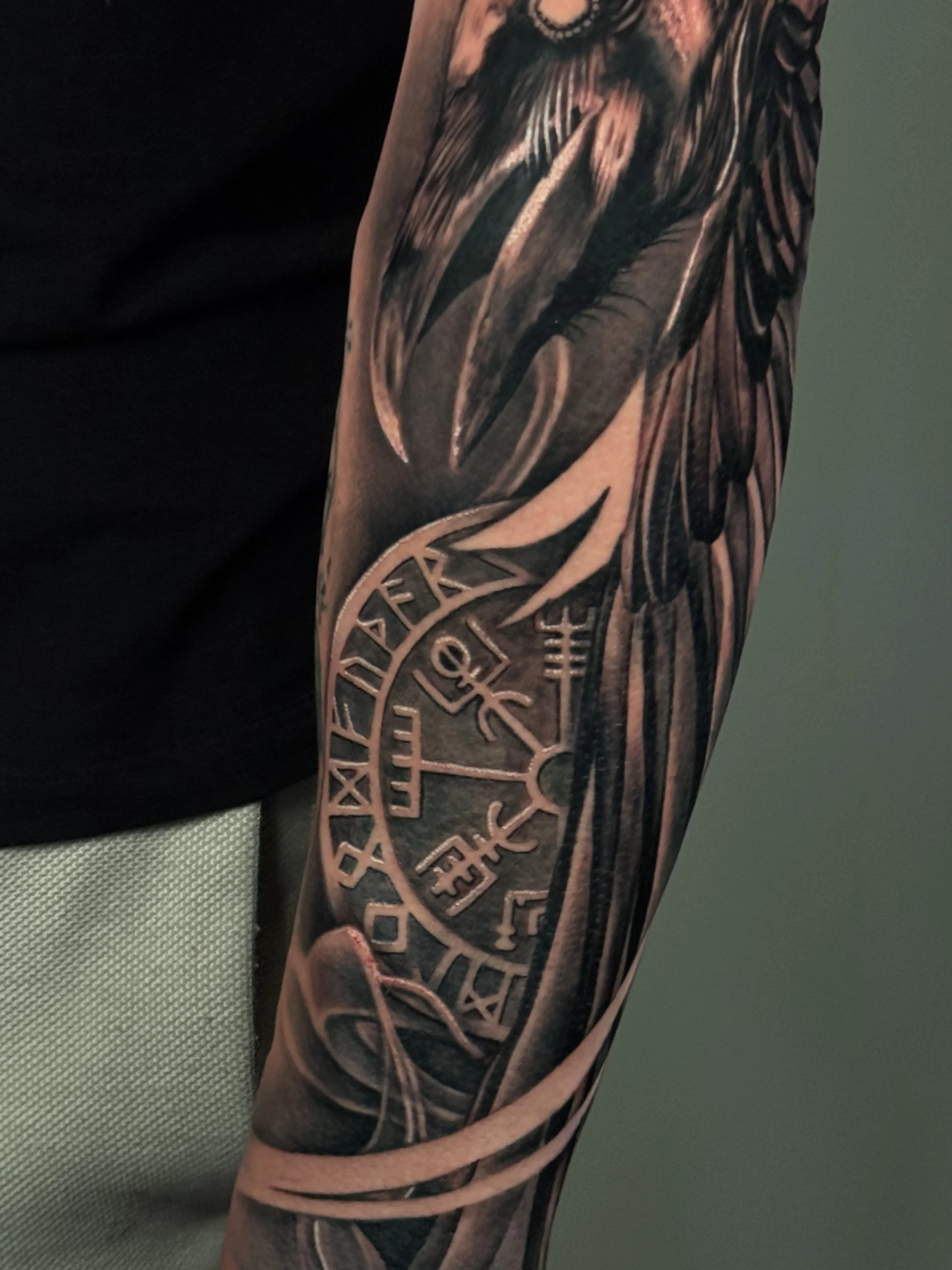 A detailed tattoo on the arm, featuring a circle with Viking or runic symbols and a raven or eagle design at the top.