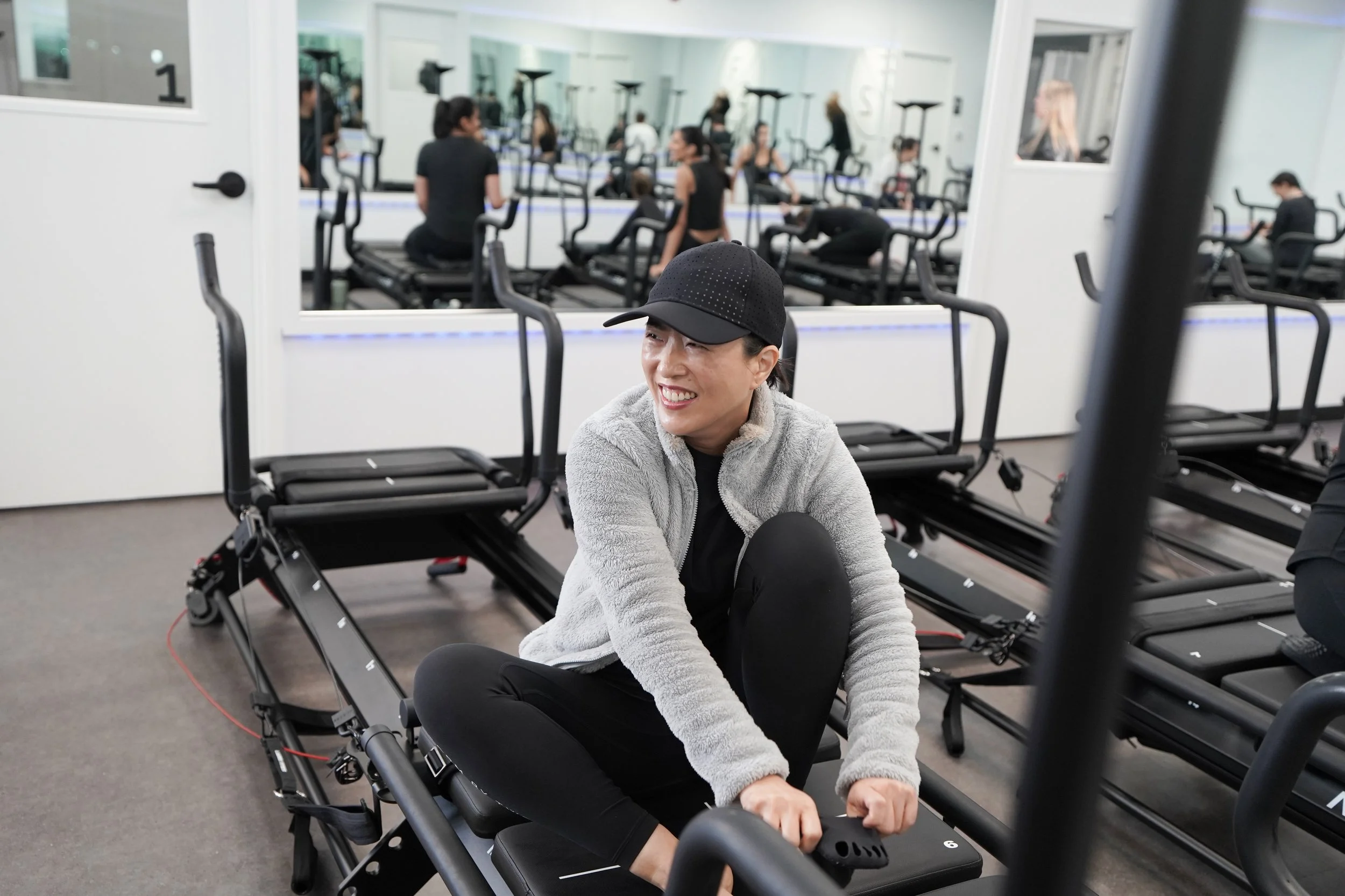 A woman sitting on a reformer Pilates machine in a gym, smiling and dressed in a black cap, gray jacket, and black leggings. Other people are working out on Pilates reformers in the background.