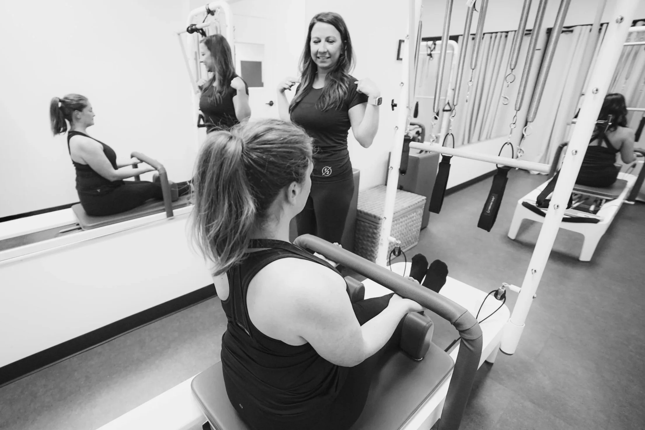 Two women perform and supervise physical exercises in a gym with fitness equipment, including a reformer and a wall-mounted exercise station.
