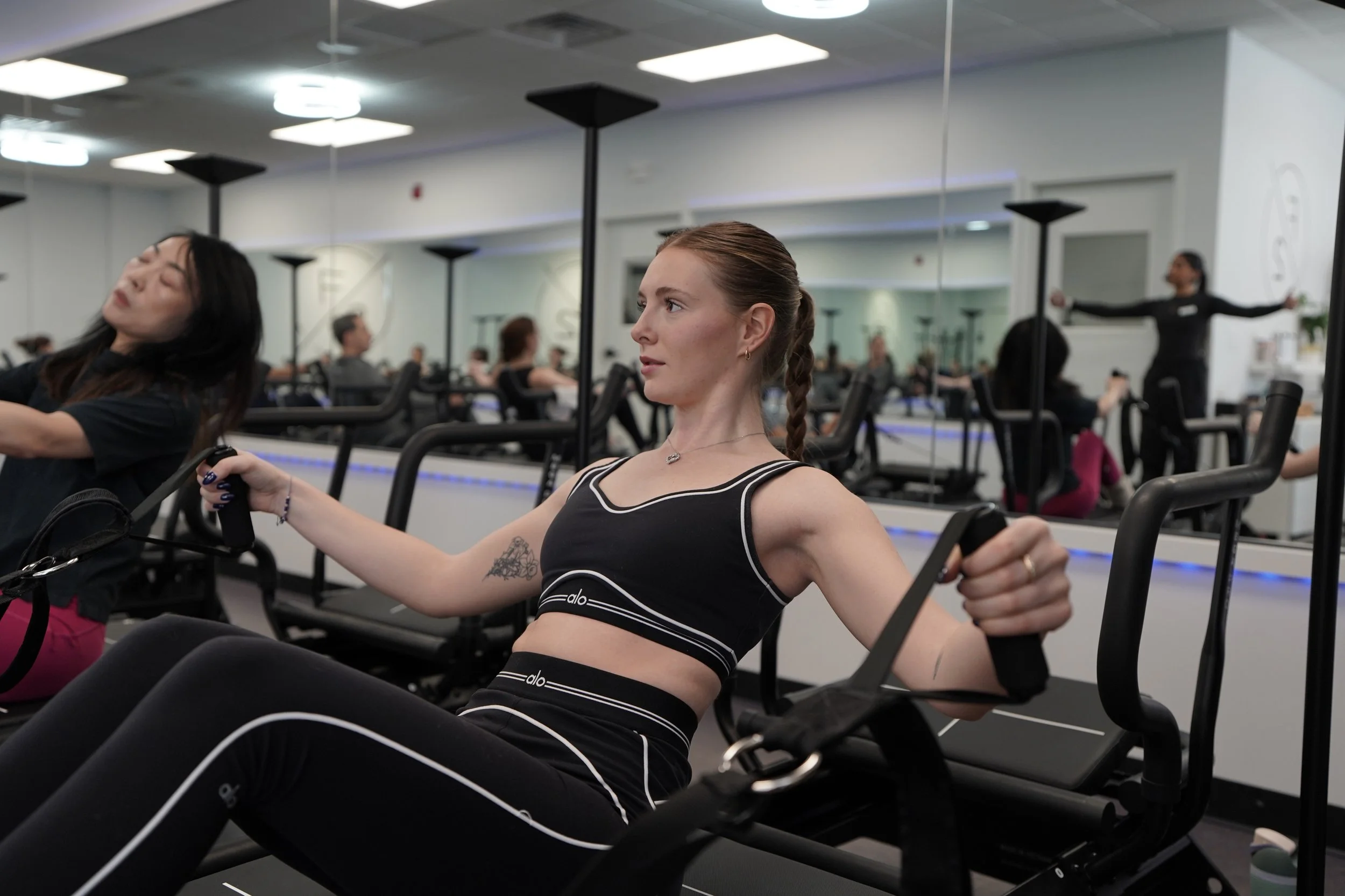 Woman exercising on a rowing machine in a gym, with mirrors and other people working out visible in the background.