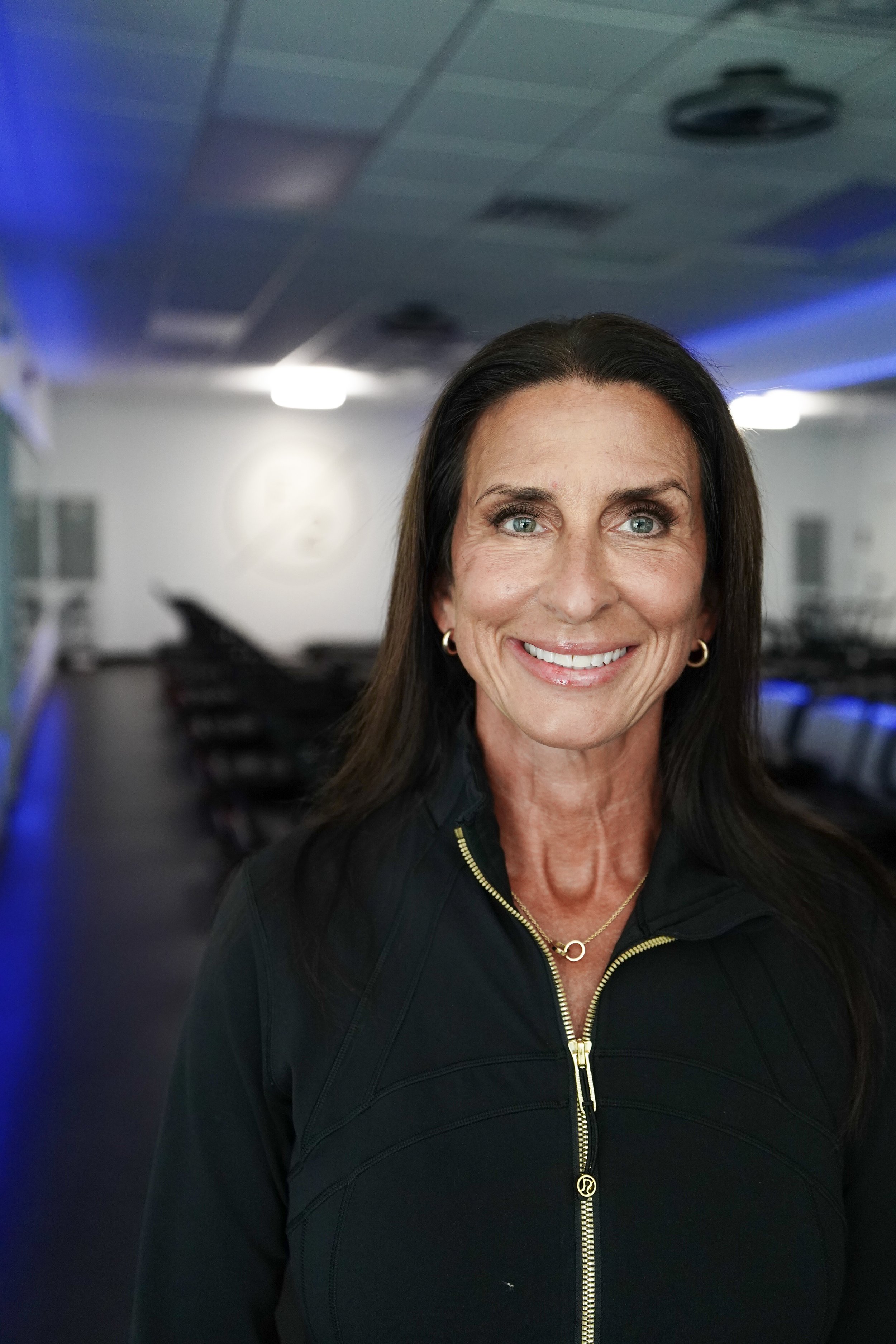 A smiling woman with long dark hair and blue eyes is standing in a room with black chairs and blue lighting accents.