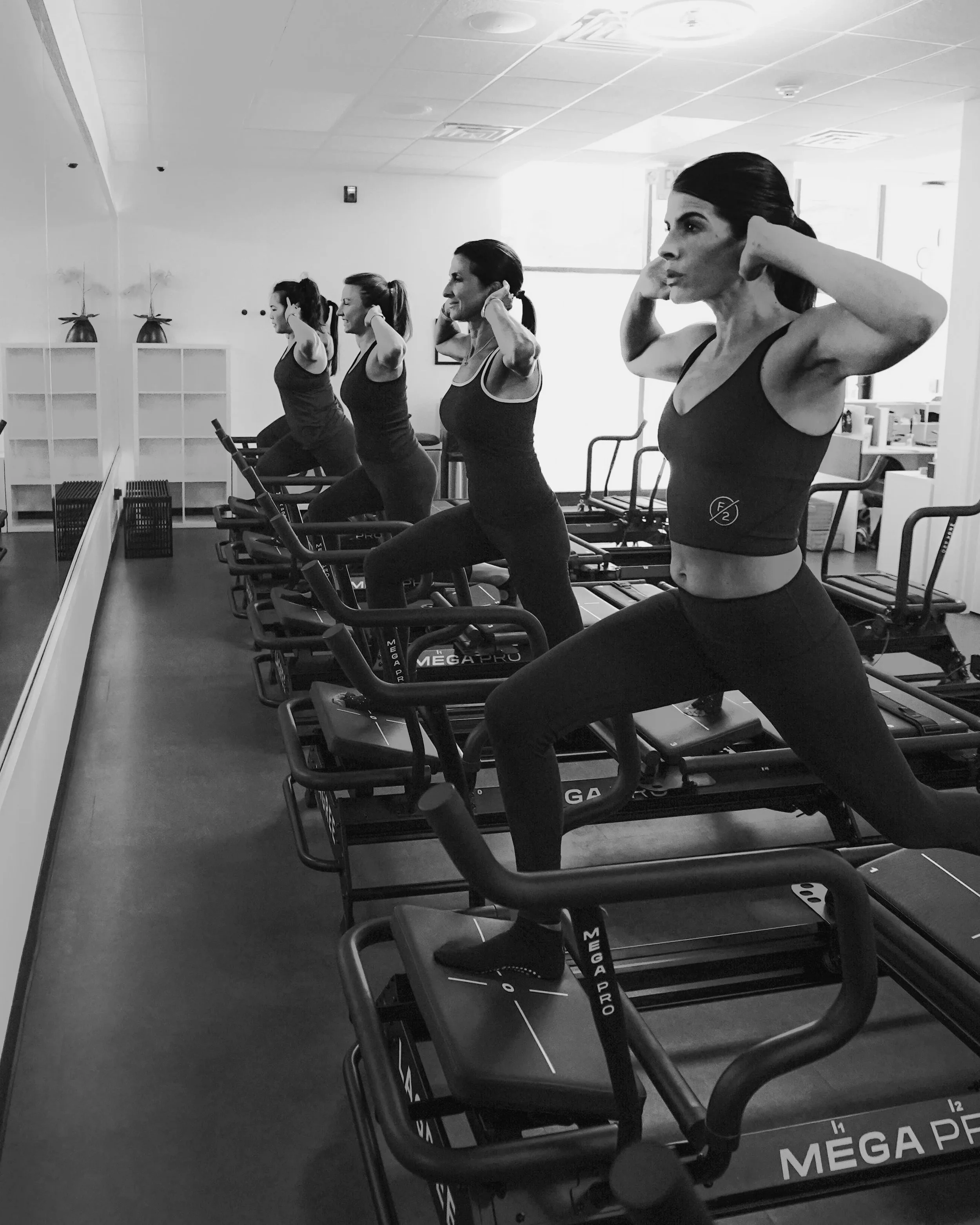 Four women in workout clothes are on Megaformer machines, performing a lunge exercise with hands behind their heads.