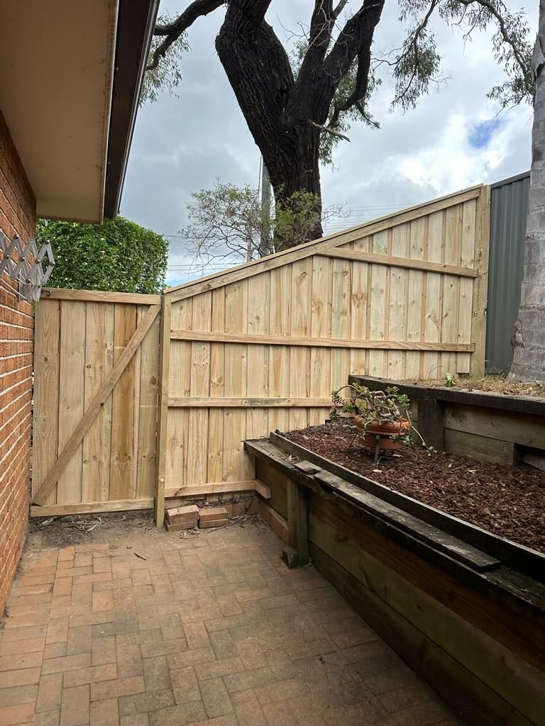 A small backyard patio with a newly built wooden fence, brick pavers on the ground, and a raised planter with a small plant. There's a large tree visible above the fence and part of the house's brick wall on the left.