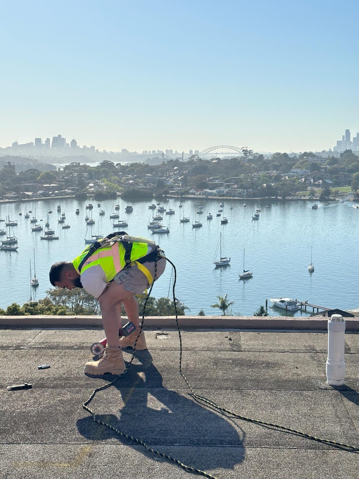 A man wearing a safety vest, shorts, and boots is working on a rooftop near a body of water filled with boats, with a city skyline and bridge in the background.
