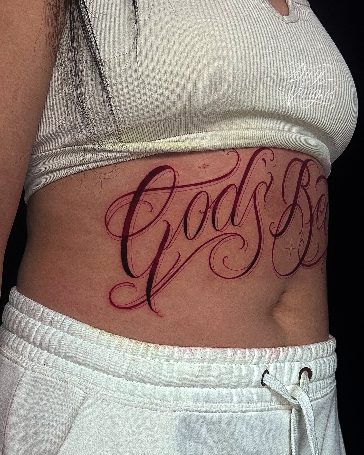 A person's side torso with a large tattoo of the word 'God's Beauty' in elegant, cursive script across the stomach.