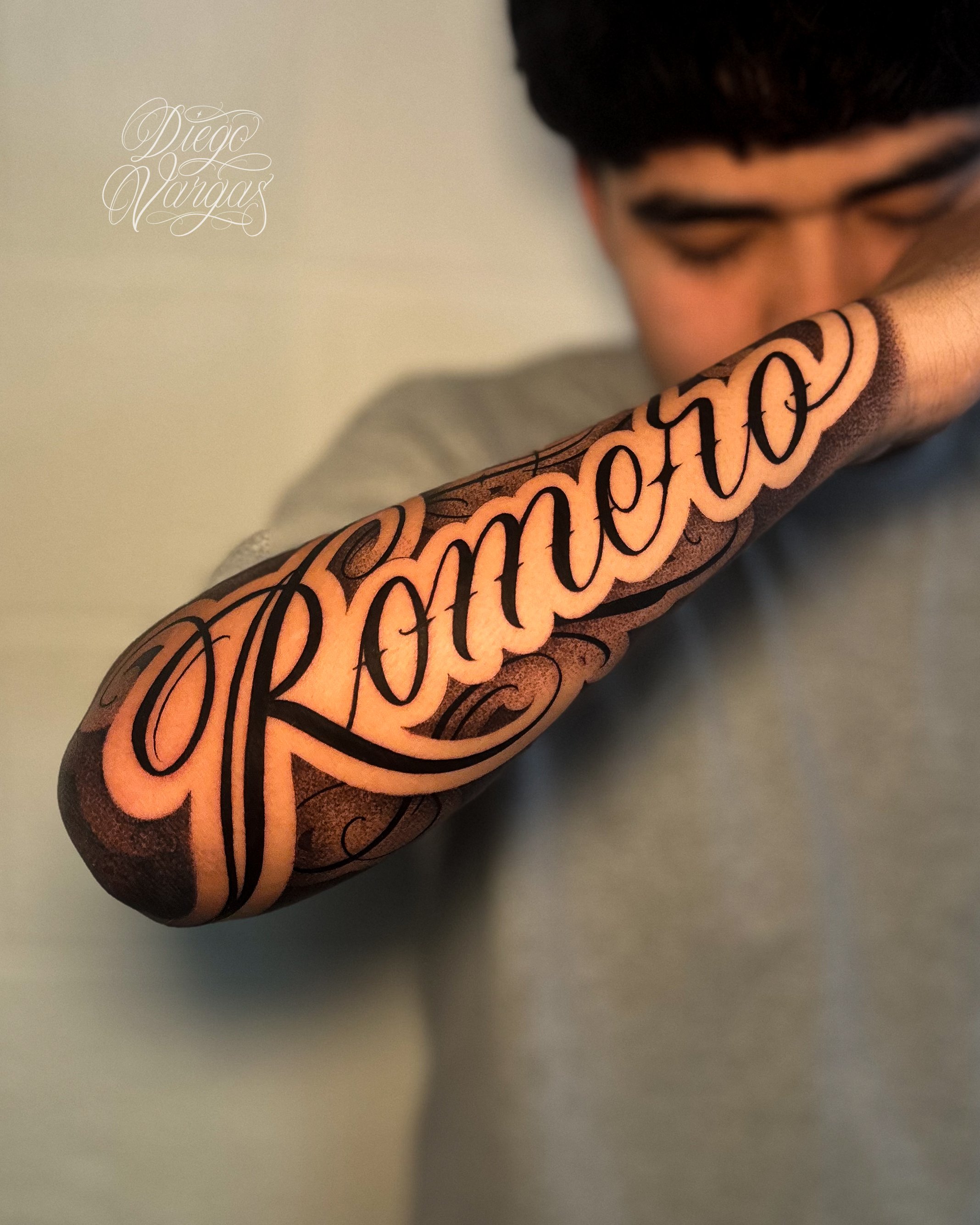 A person with a tattooed arm that reads 'Romero' in bold, cursive script, with some intricate decorative elements around the text.