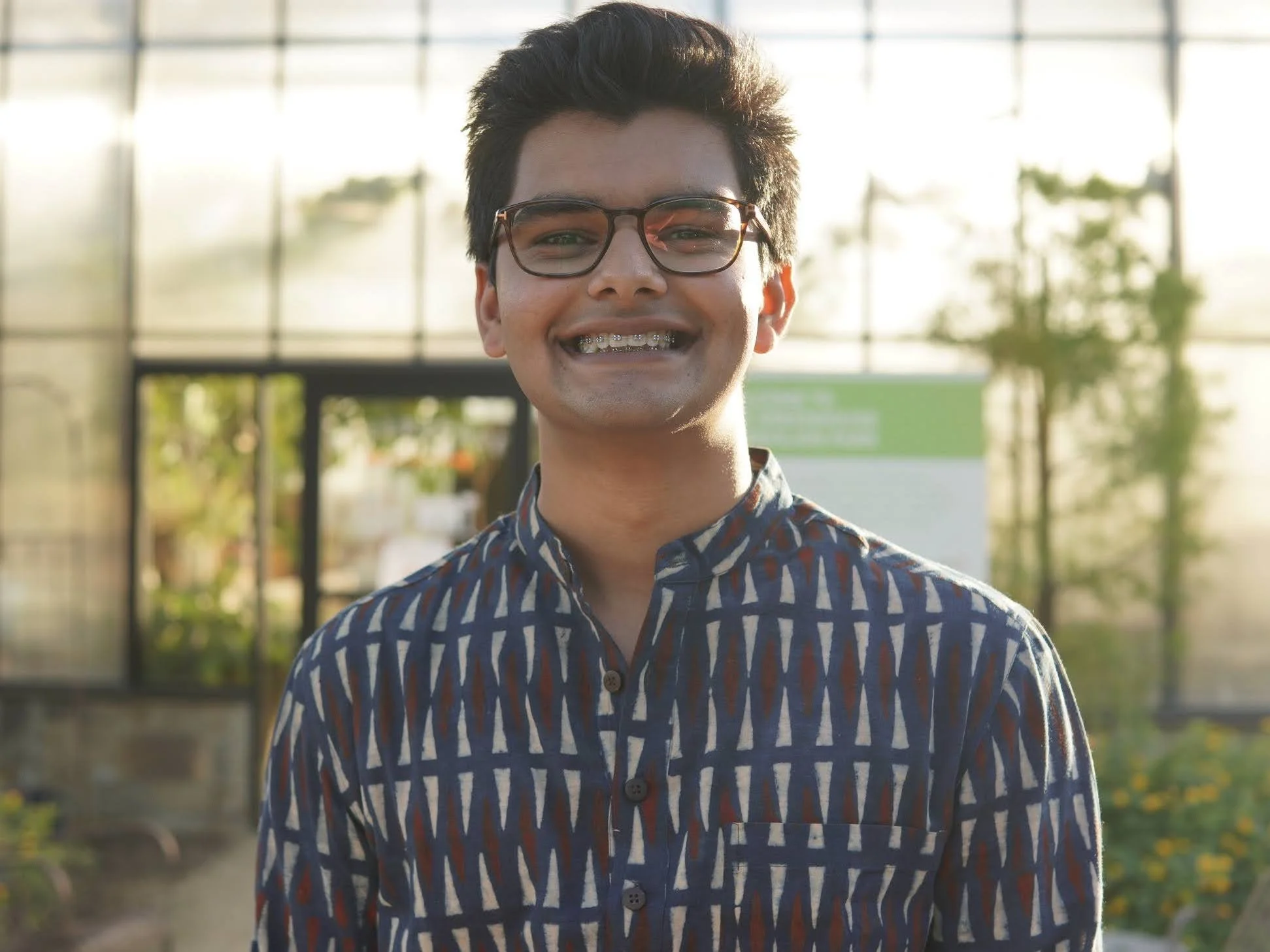 Smiling person wearing glasses and a patterned shirt standing in front of a greenhouse with plants in the background.