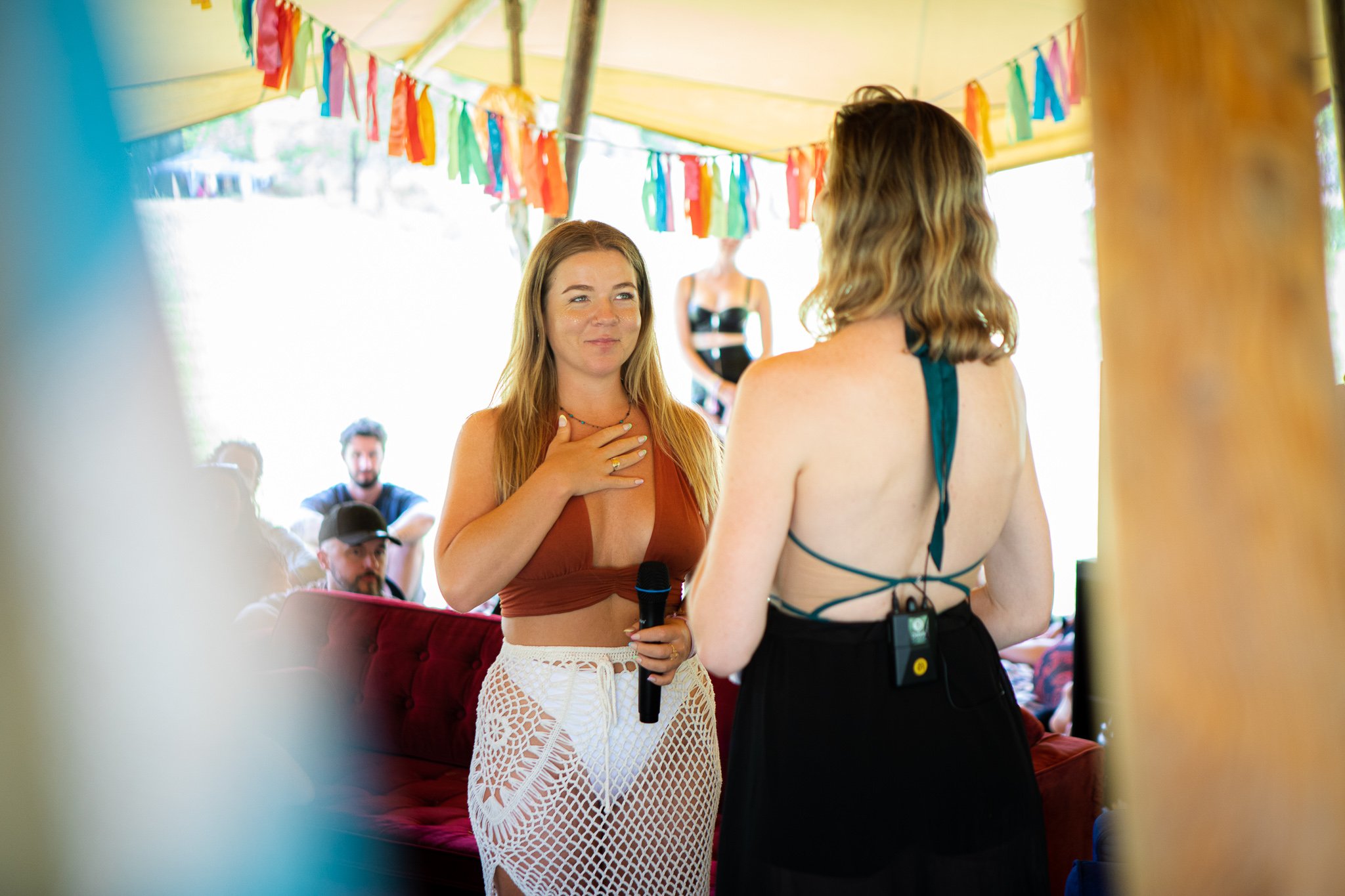 Two women are speaking to each other indoors during a party or celebration, with colorful decorations hanging overhead and a group of people in the background.