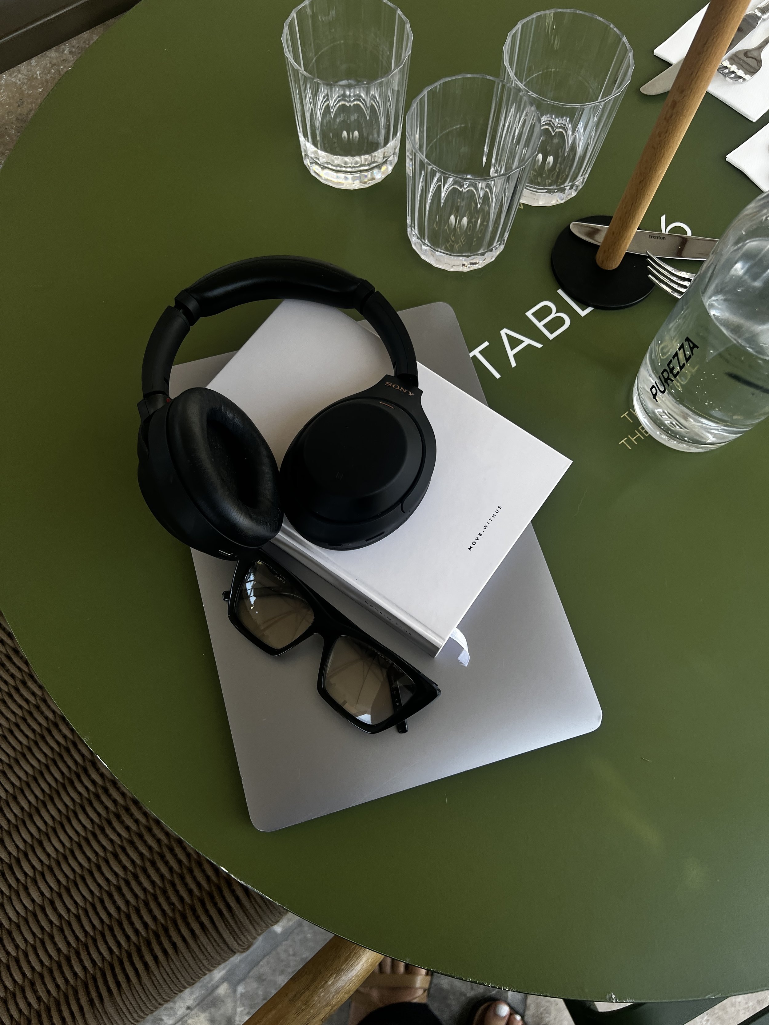 A green round table with three empty drinking glasses, a black-headphone, black sunglasses, a white box, a bottle of water, a fork, a knife, and a wooden stand with a black base.