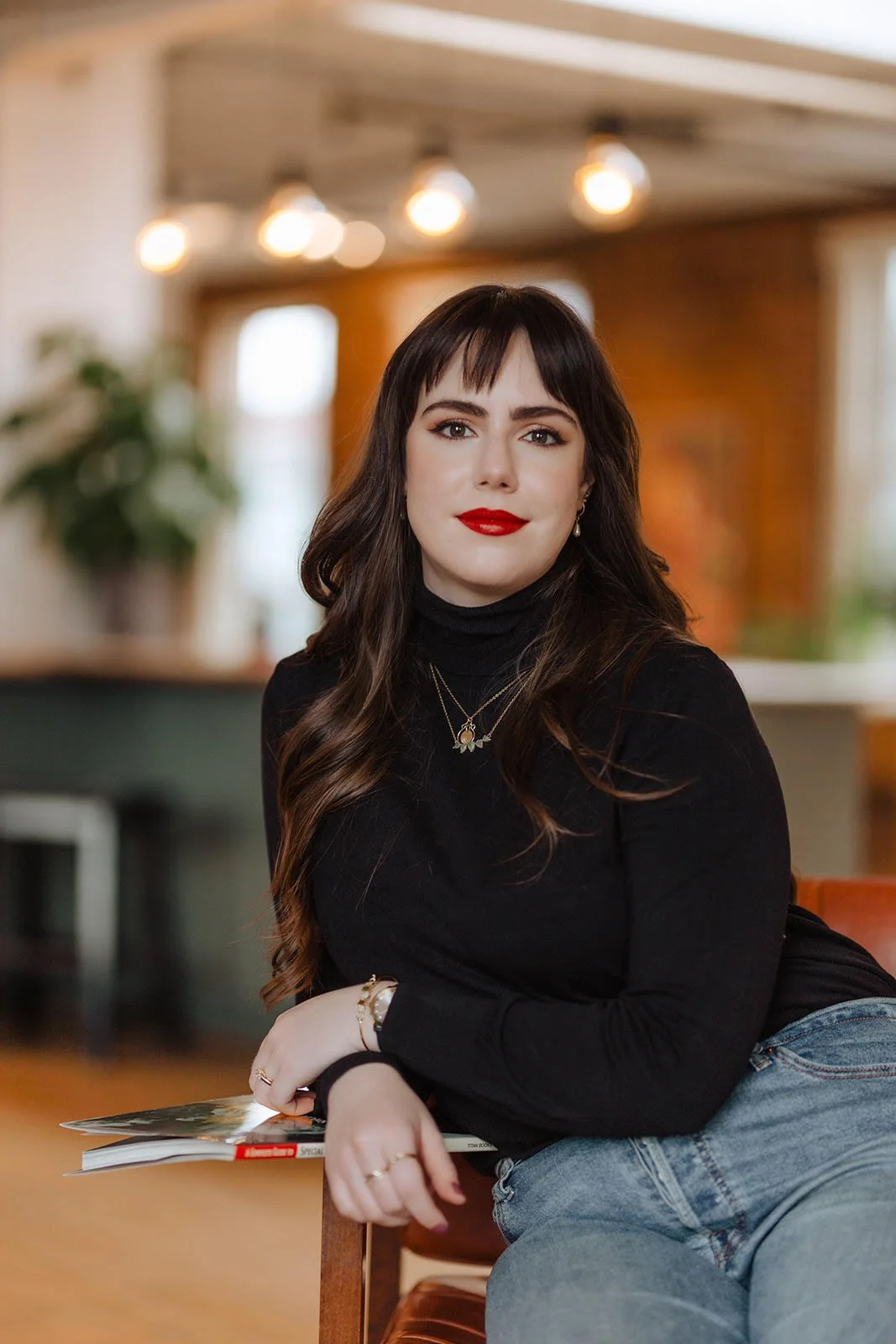 A young woman with long wavy brown hair, wearing a black turtleneck and gold jewelry, sitting in a cozy, well-lit indoor space with warm lighting and a blurred background.