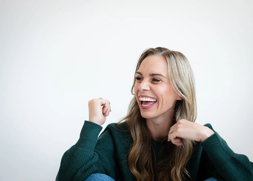 A woman with long blonde hair wearing a dark green sweater, smiling enthusiastically, with her fists raised in celebration or excitement.