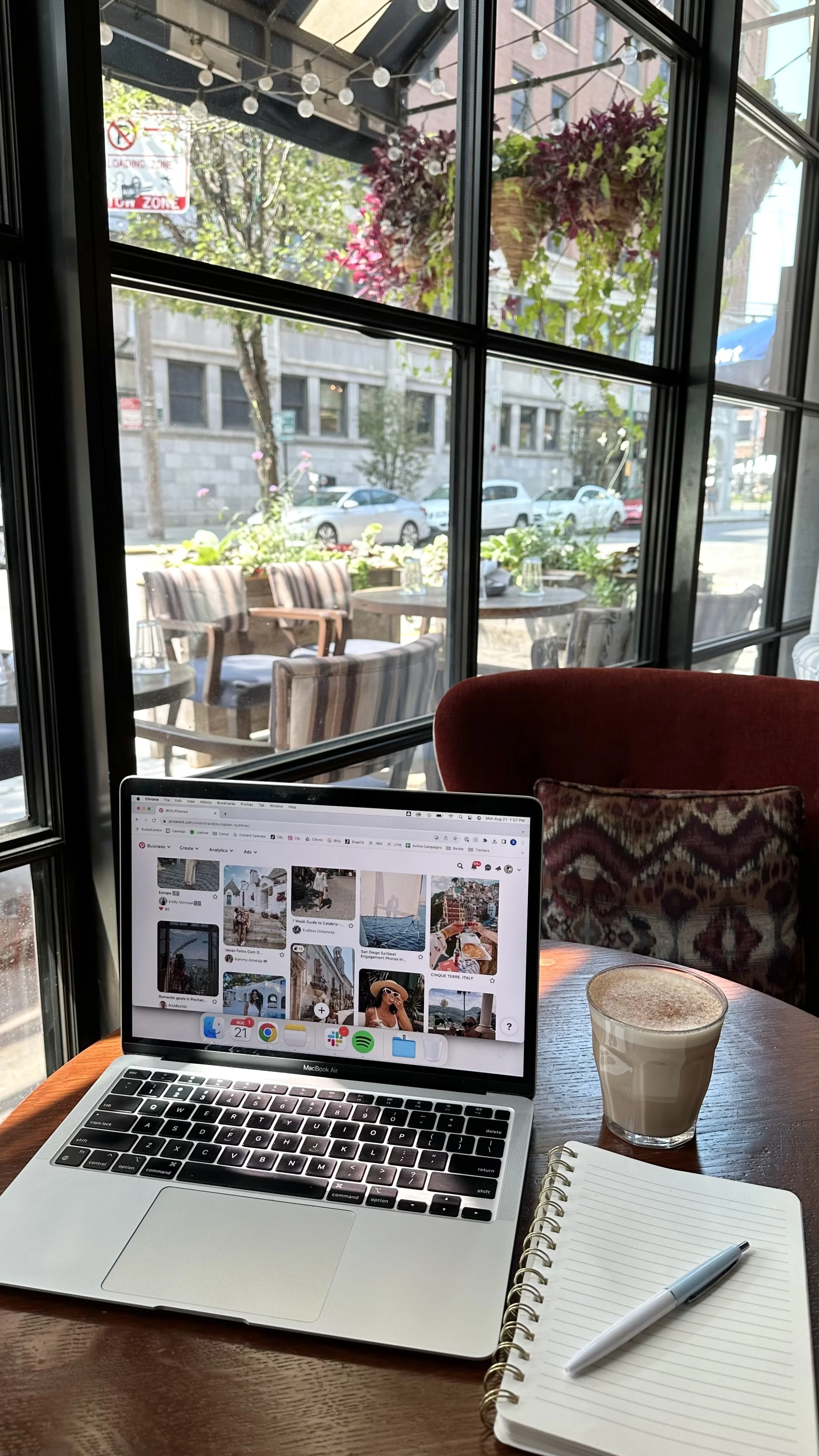 A cozy cafe scene with a MacBook Air displaying a Pinterest feed on a round wooden table. The table also has a notebook and pen, and a glass of coffee. Outside the window, there are outdoor seating with chairs and tables, plants, and a street with parked cars.