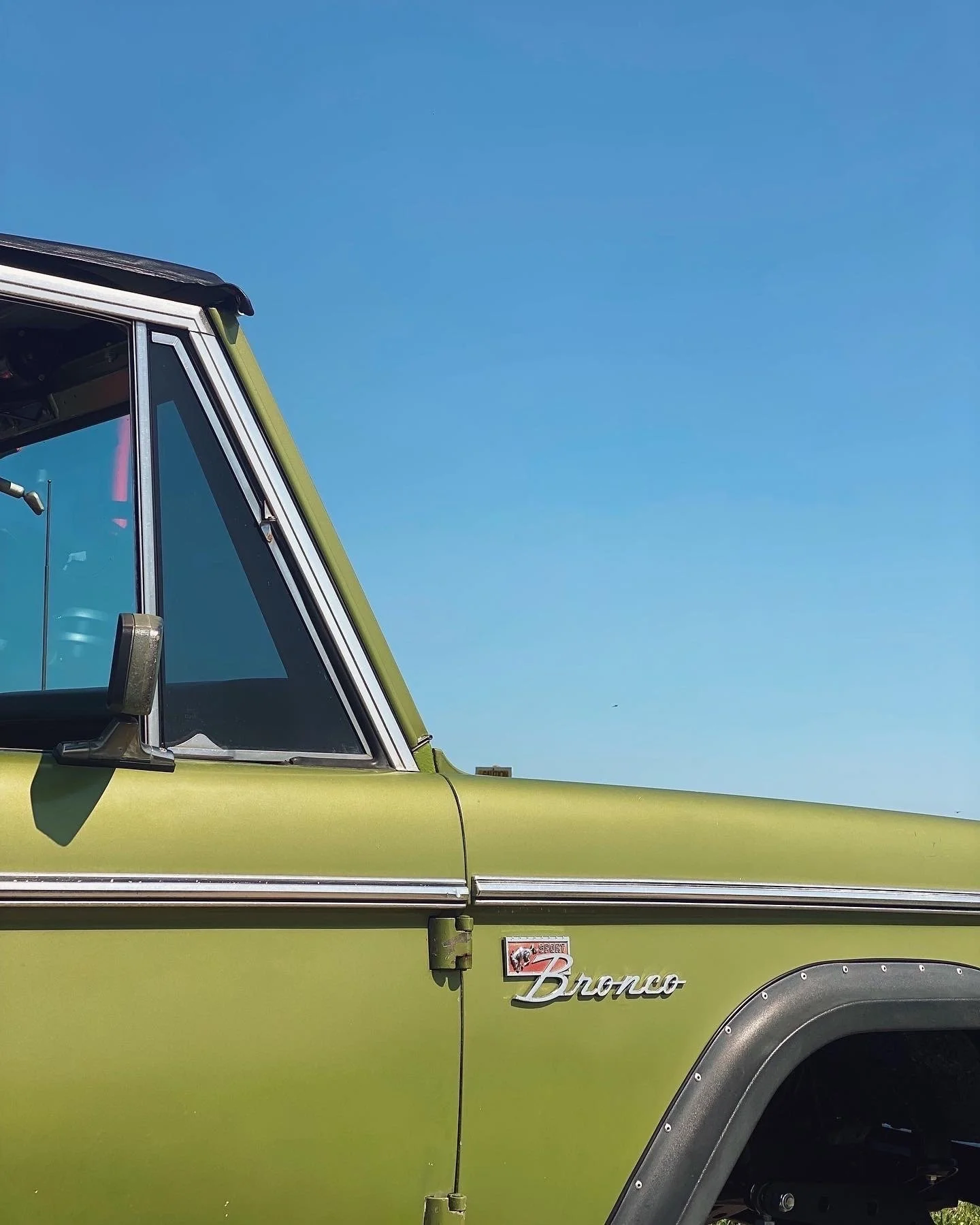 Close-up of the front side of a vintage green Ford Bronco showing the windshield, door, and logo under a clear blue sky.