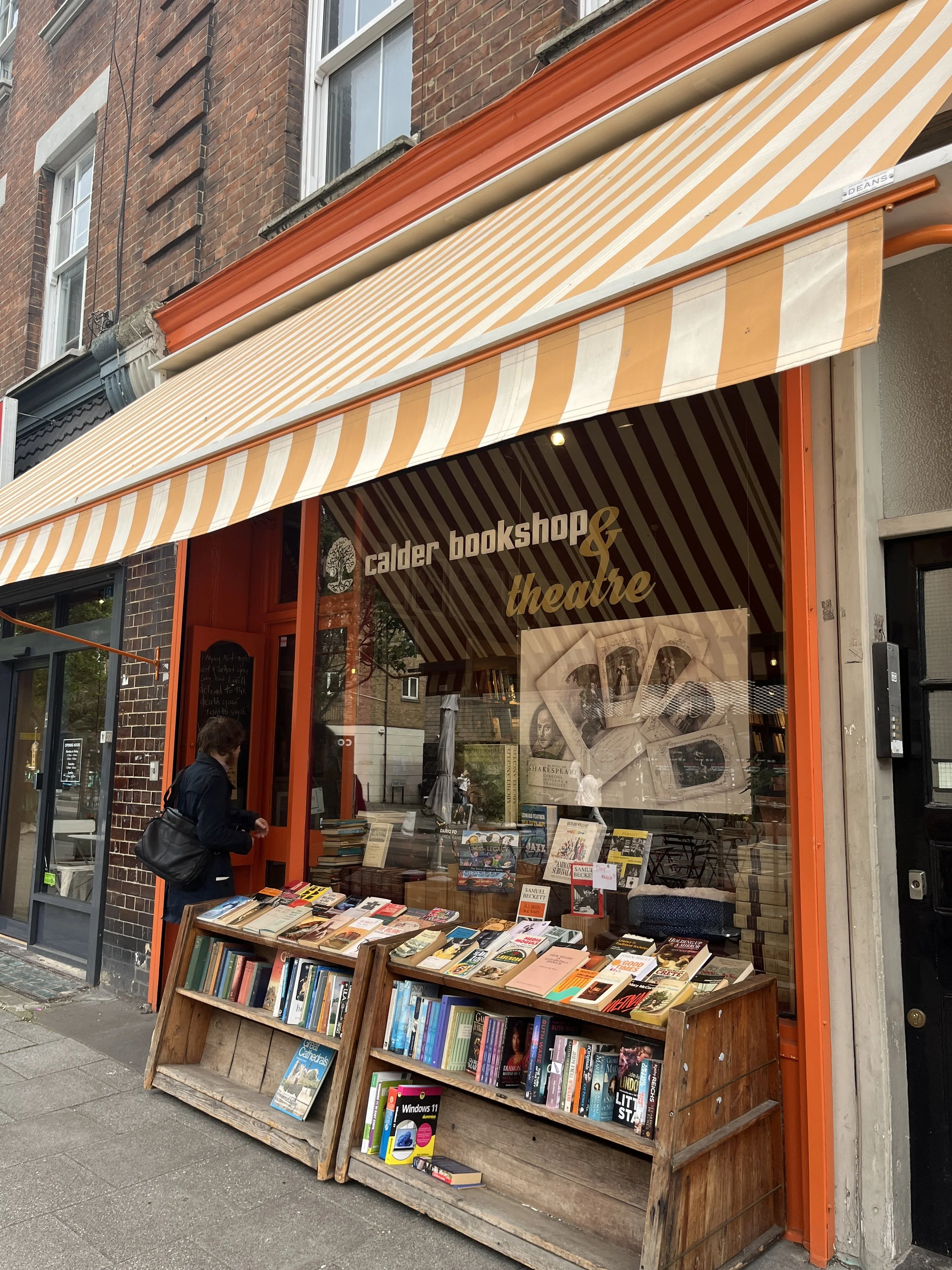 A street view showing the storefront of Calder Bookshop & Theatre with books displayed outside under an cream and orange striped awning. A person is browsing the books.