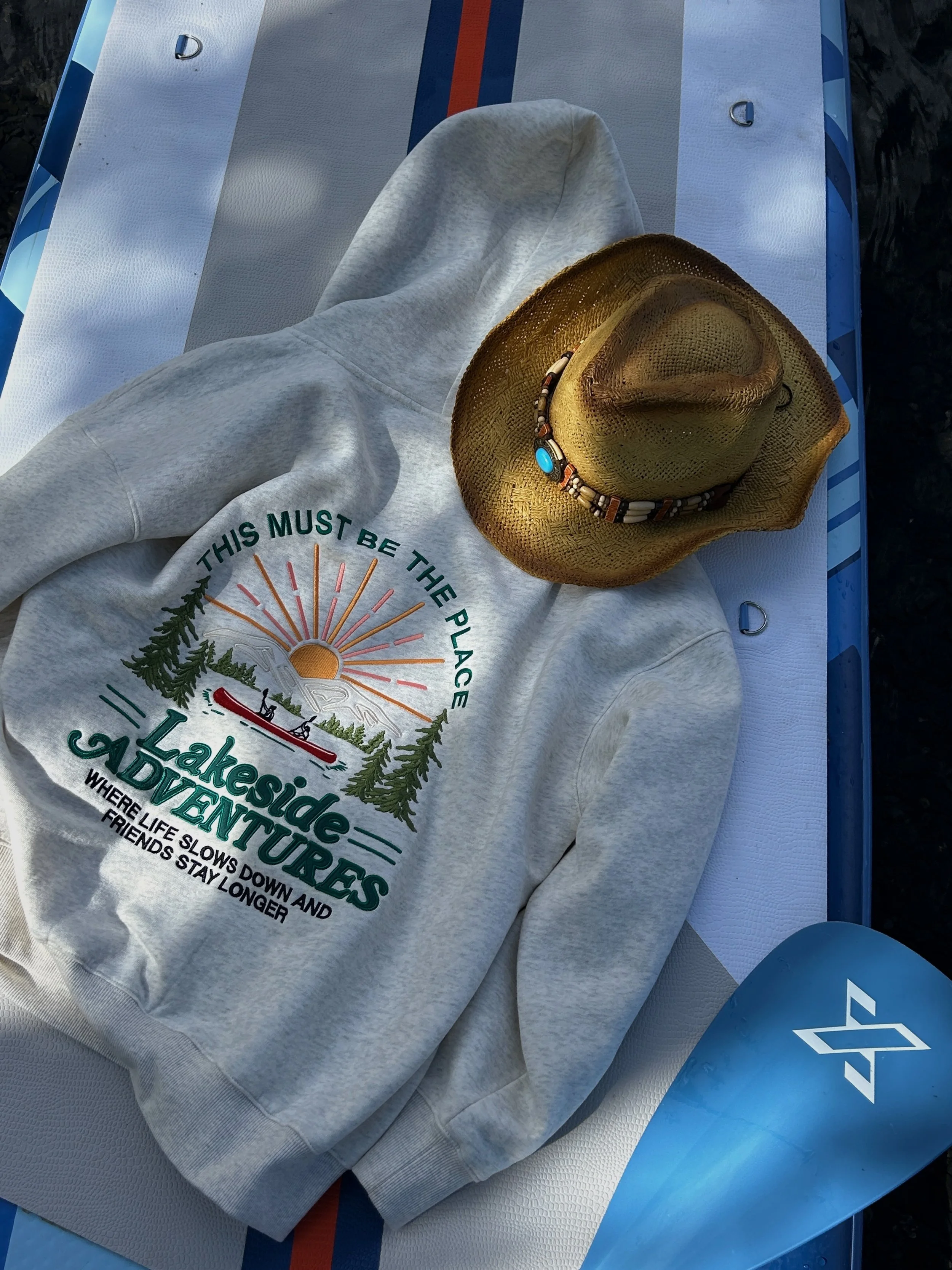 A white T-shirt with a colorful logo reading "Lakeside Adventures" lying on a white surfboard with a blue stripe and two D-rings. A straw hat with a beaded band rests on top of the T-shirt. Part of a blue paddle with a logo is visible in the corner of the image.