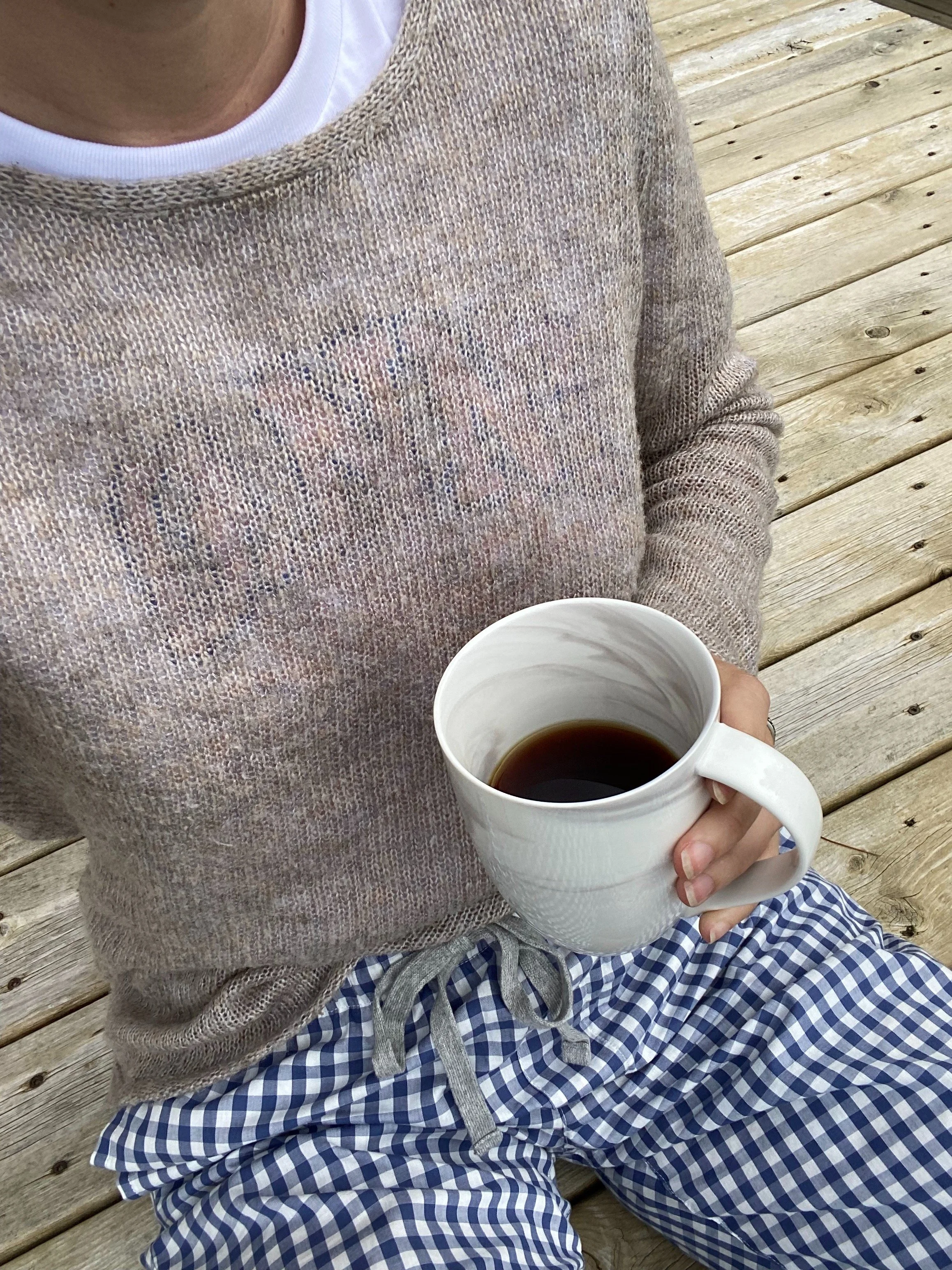 A person holding a white mug of coffee while sitting on a wooden deck, wearing a beige knit sweater over a white shirt and blue checkered pajama pants.