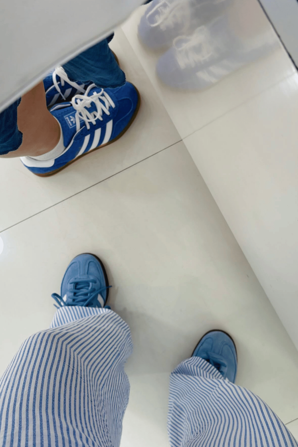 Person in striped shorts and blue shoes standing on a white tiled floor, with another person wearing blue sneakers and blue jeans nearby.