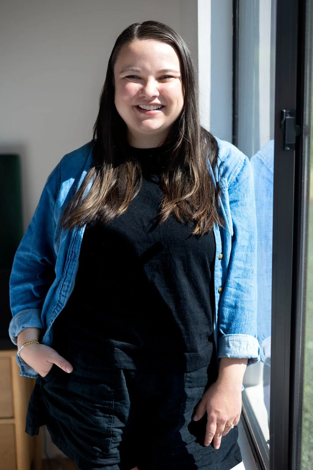 A young woman with shoulder-length brown hair, wearing a black t-shirt and a blue denim jacket, standing next to a window and smiling.