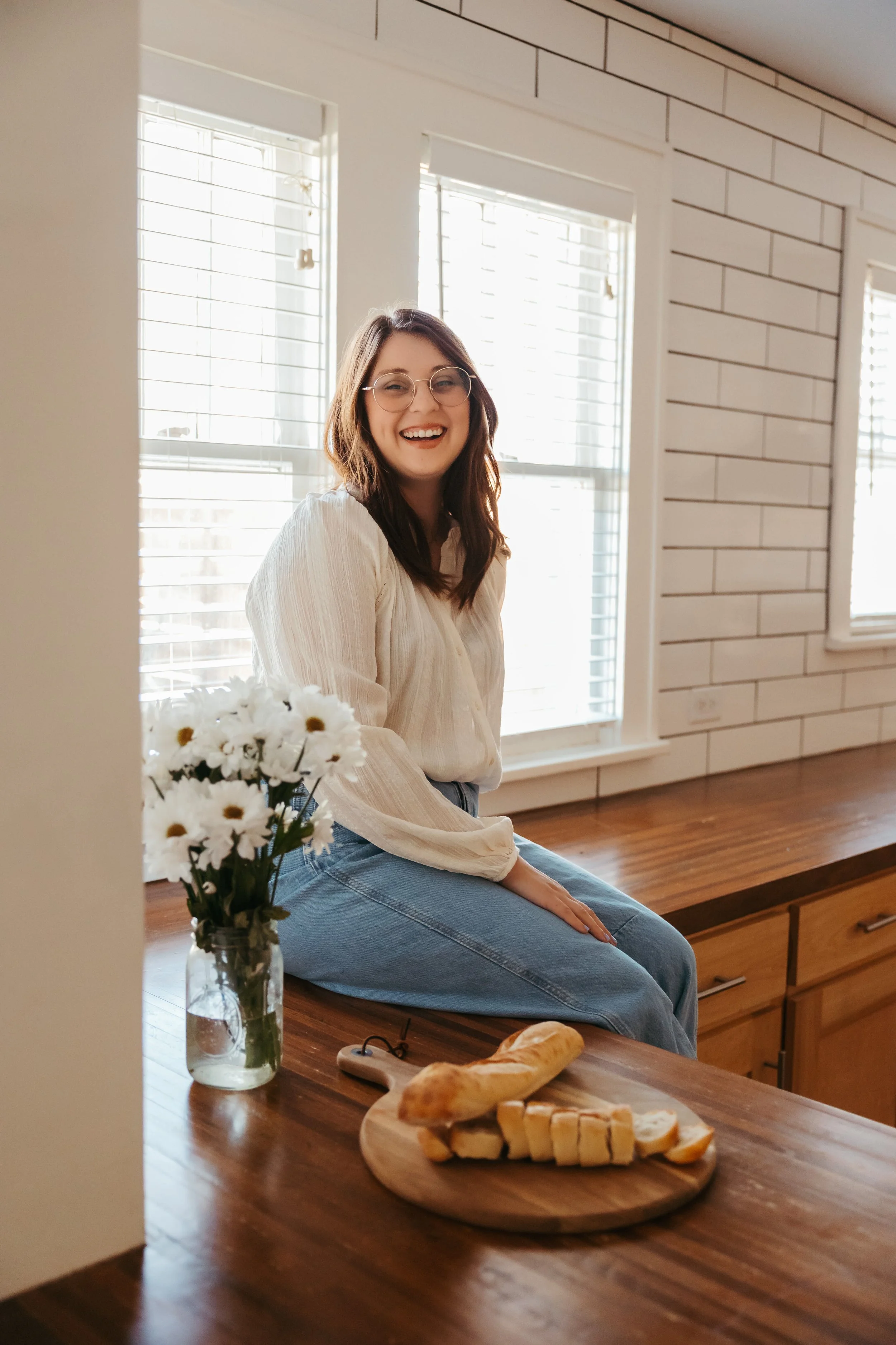 A young woman with glasses and a cream blouse sitting on a kitchen counter, smiling, with a vase of daisies and a wooden cutting board with bread in front of her, in a bright kitchen with large windows and white brick walls.