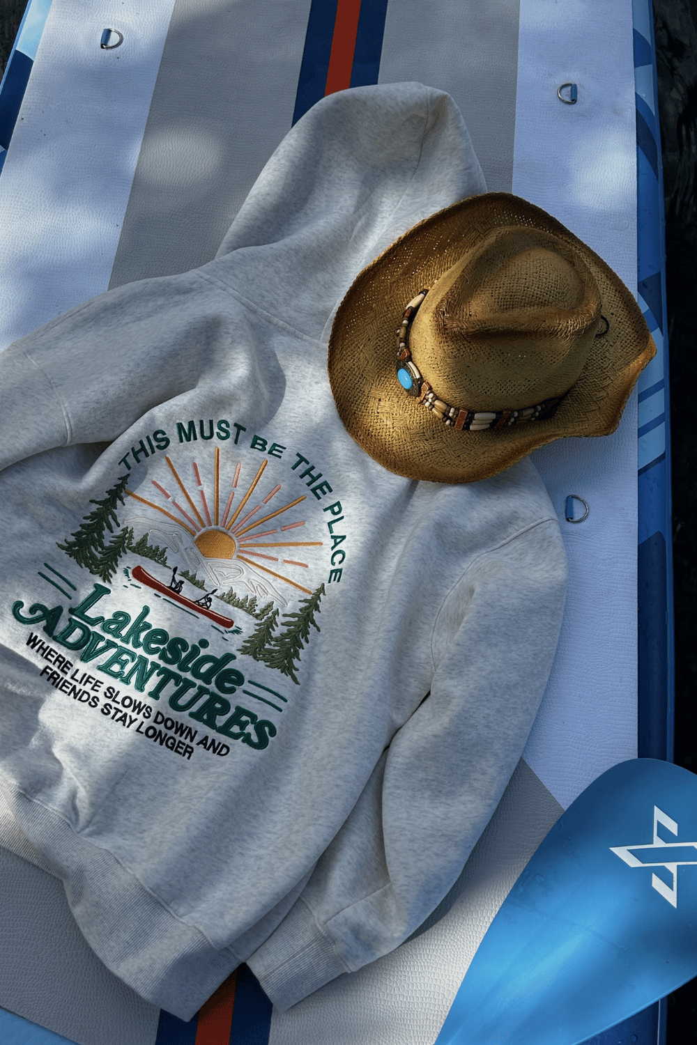 A beige cowboy hat with a beaded band and a gray hoodie with a lakeside adventure design and sunset, lying on a white and blue paddleboard.