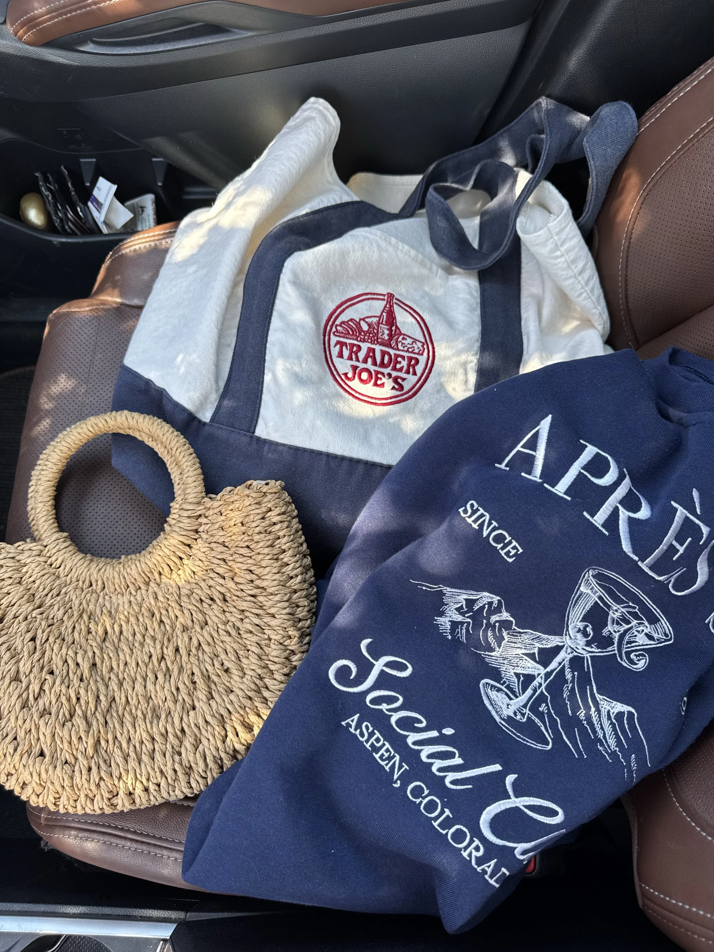 Inside a car front passenger seat with various bags and items including a blue tote bag with white embroidery, a tan woven straw purse, and a Trader Joe's shopping bag, with some personal items stored in the back seat