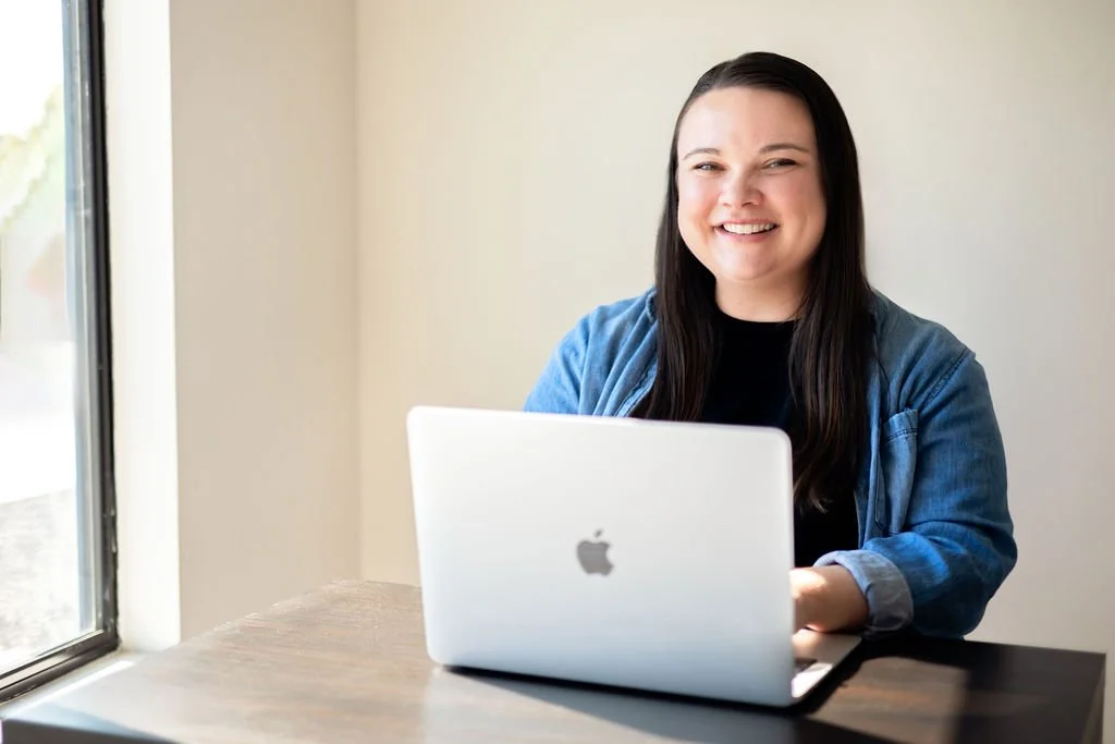 A smiling woman with long dark hair sitting at a wooden table with an open MacBook near a window.