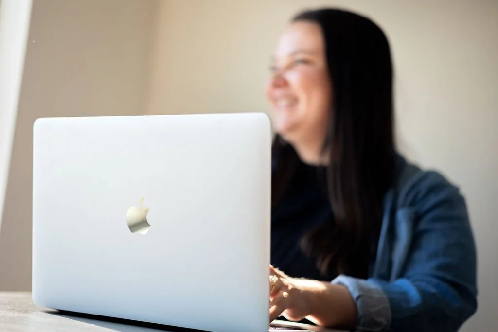 A woman with long dark hair smiling while using a silver MacBook laptop on a desk.