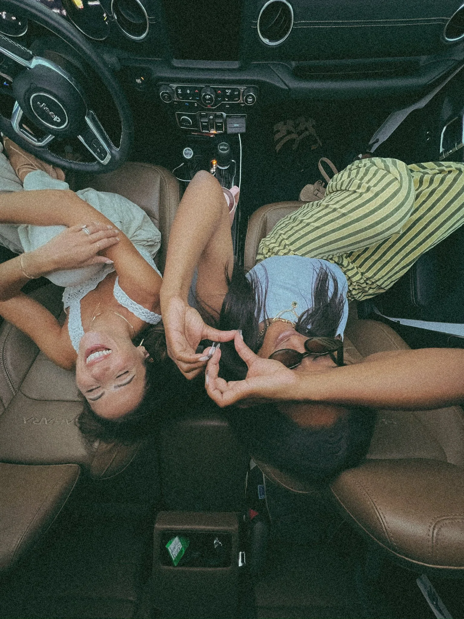 Interior of a car viewed from the backseat, showing the dashboard, steering wheel, and a person with long dark hair wearing a light-colored top and striped pants sitting in the front passenger seat, with accessories on her wrist and glasses on her head.