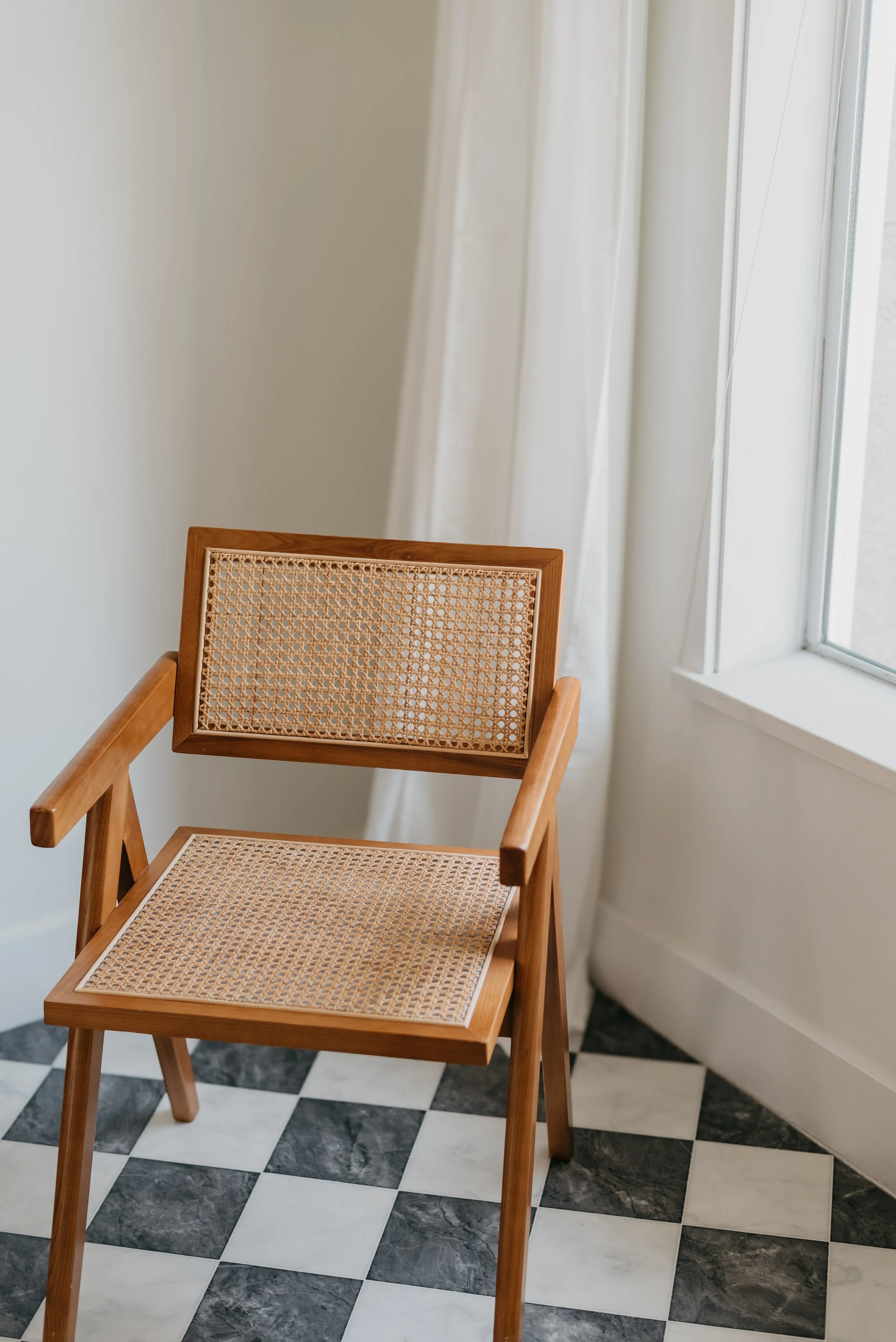 Wooden chair with woven cane seat and back, positioned near a window with white curtains in a room with black and white checkered marble floor.