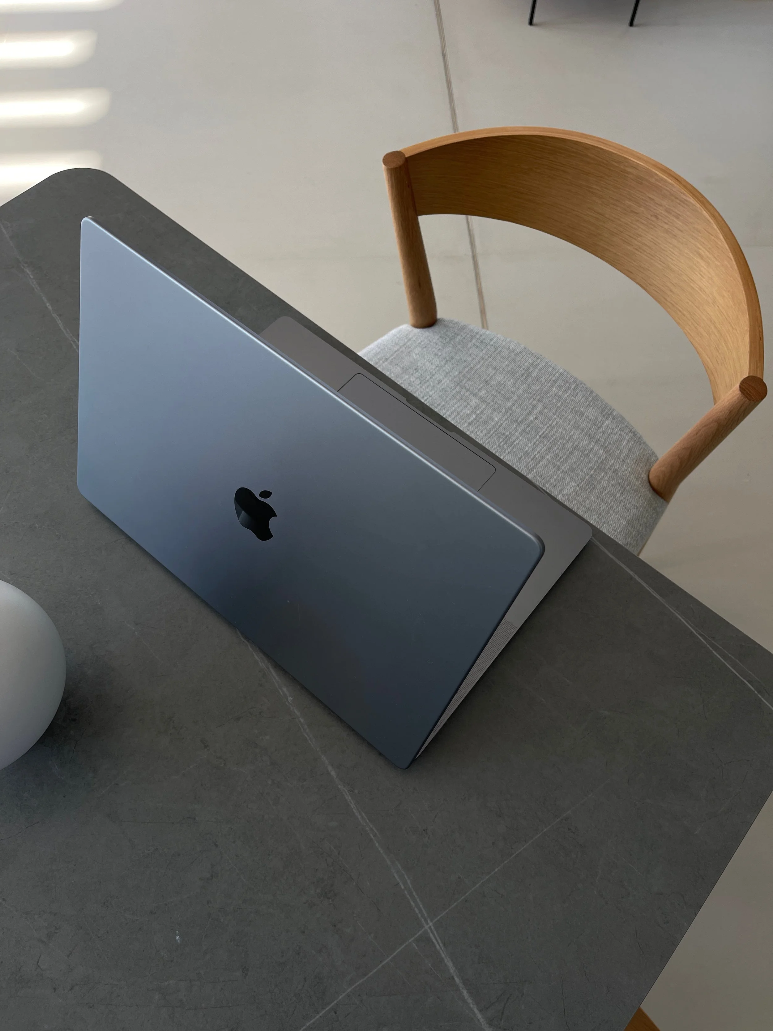 A silver Apple MacBook laptop resting on a grey stone table, green upholstered chair with wooden armrest nearby, and a grey ball on the floor in a modern, minimalistic room.