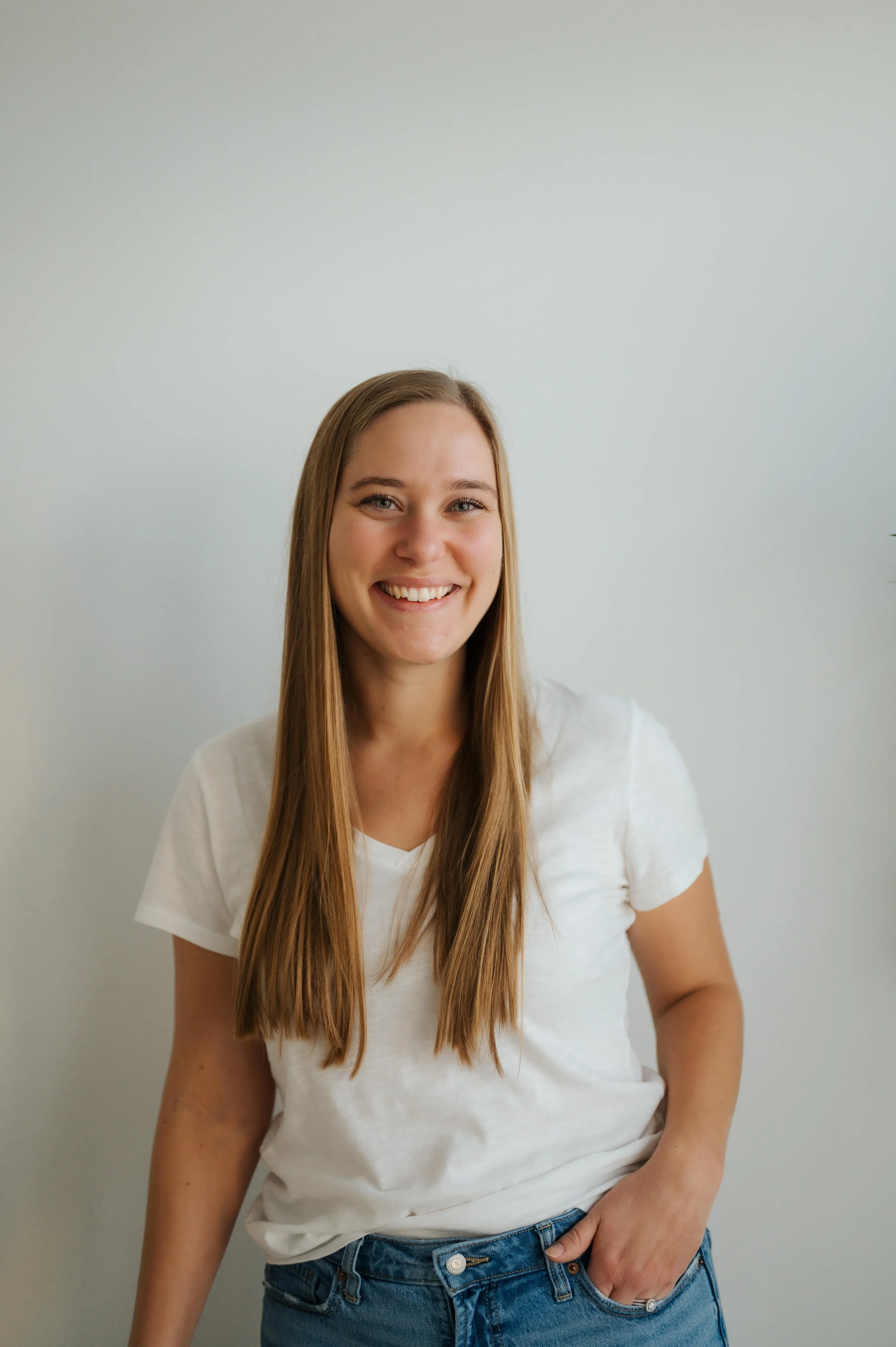 A young woman with long blonde hair, wearing a white T-shirt and jeans, smiling and standing against a plain white wall.