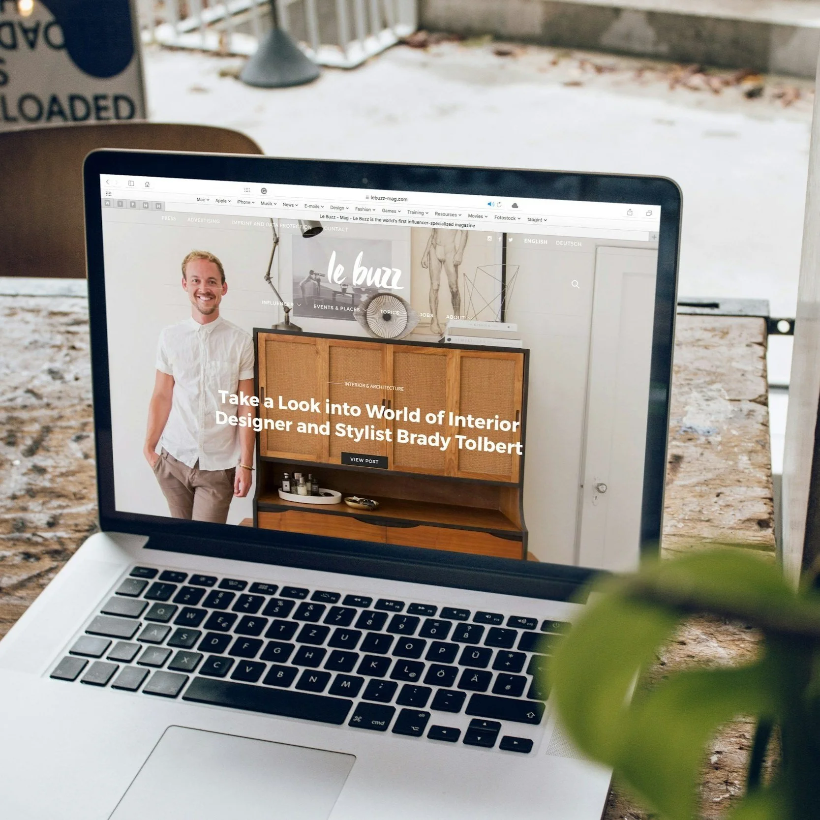 Laptop op een houten tafel toont een website over interieurdesign en stylisten Brady Tolbert, met een man die glimlachend in de afbeelding op de site staat.
