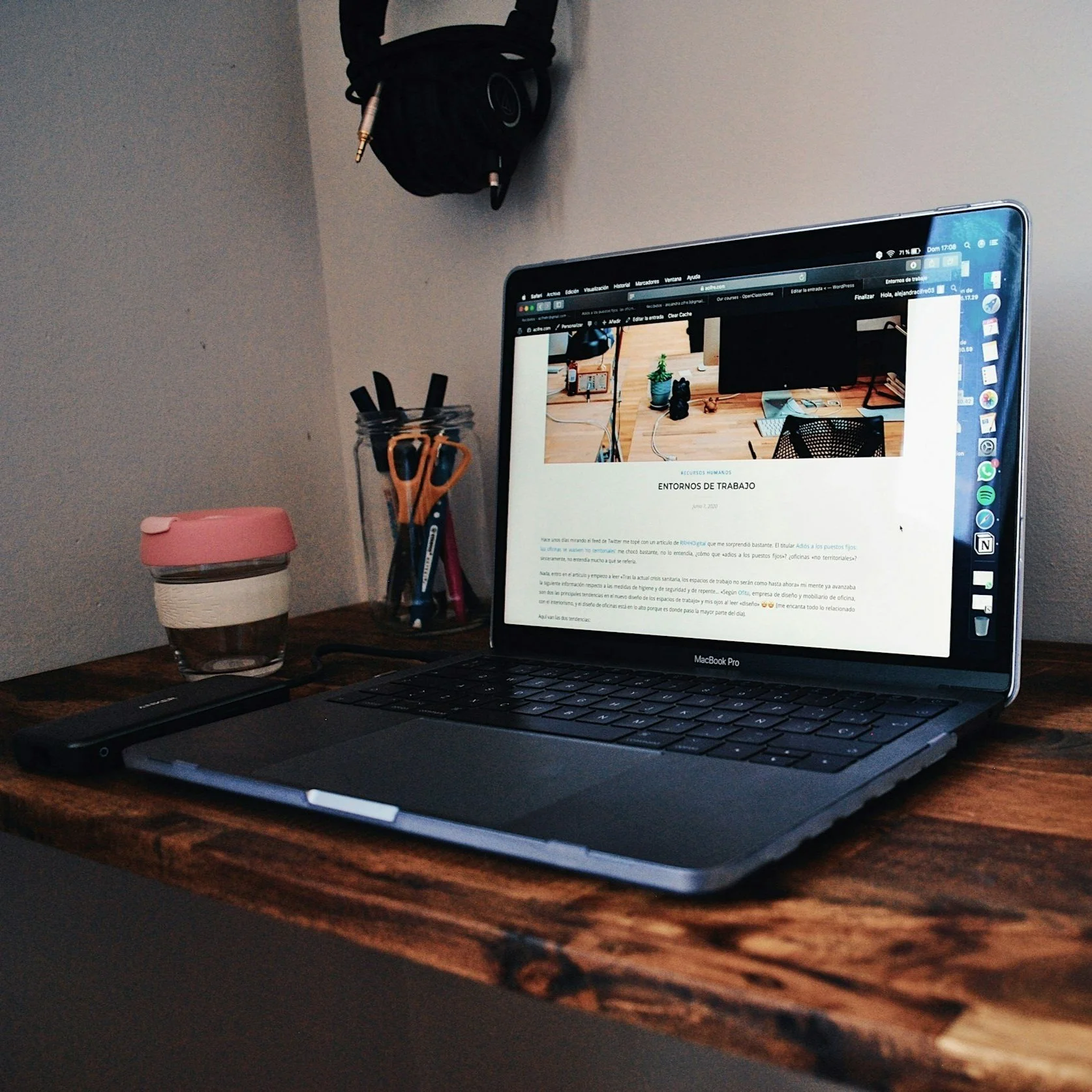 Een open MacBook Pro op een houten bureau met een website over werkplekken op het scherm. Op de bureaublad staan pennen en scharen in een glazen pot, een zwart apparaat, en een pot met een roze deksel. Aan de muur hangt een koptelefoon.