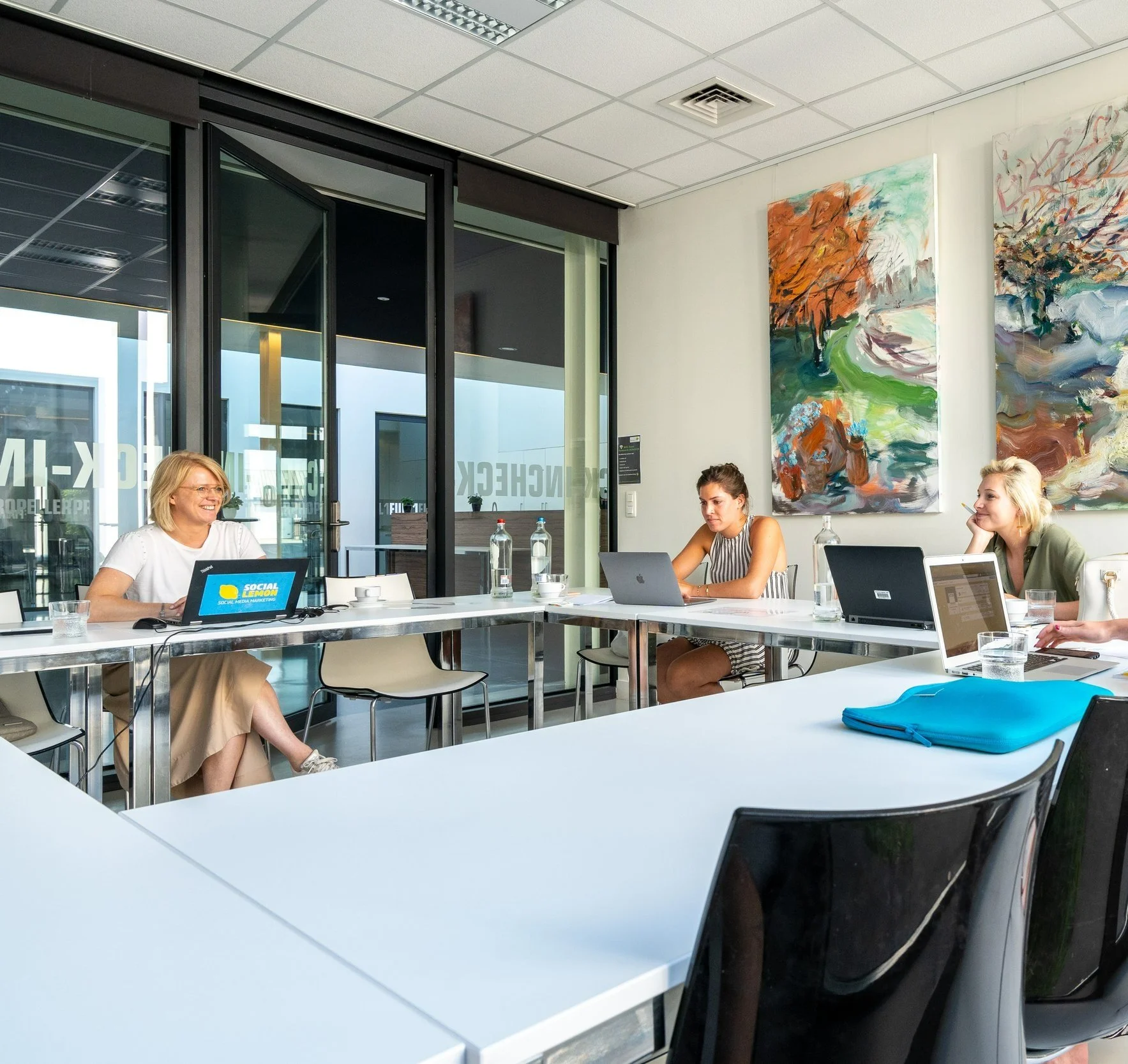 Vrouwen zitten in een vergaderruimte met laptops, waterflessen, en kleurrijke kunstwerken aan de muur.
