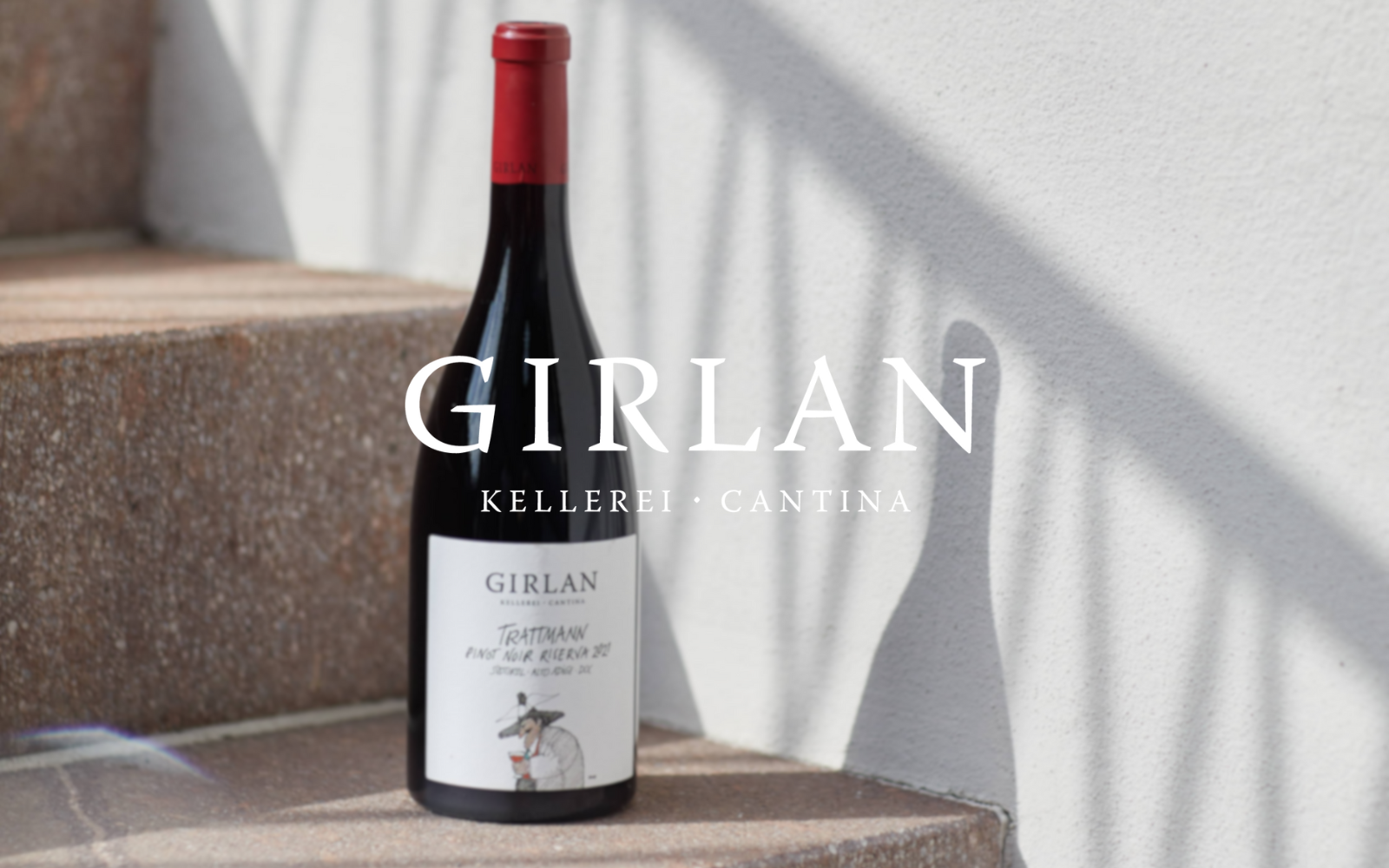 A bottle of Girlan Trattmann First Noir Riserva red wine on a stone step with shadows cast on a white wall behind it.