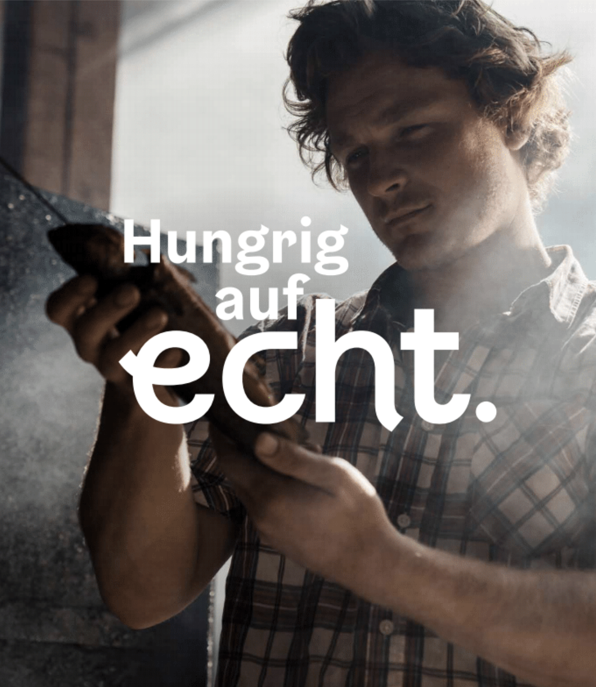 A person with brown curly hair wearing a plaid shirt checking his phone while leaning against a dark stone wall. The background is bright and appears to be outdoors. Overlaid text says, 'Hungrig auf echt.'