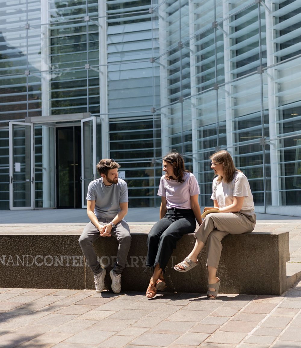 L'immagine mostra tre membri del team seduti fuori dal museo contemporaneo Museion che parlano tra loro, seduti su una panchina di pietra.