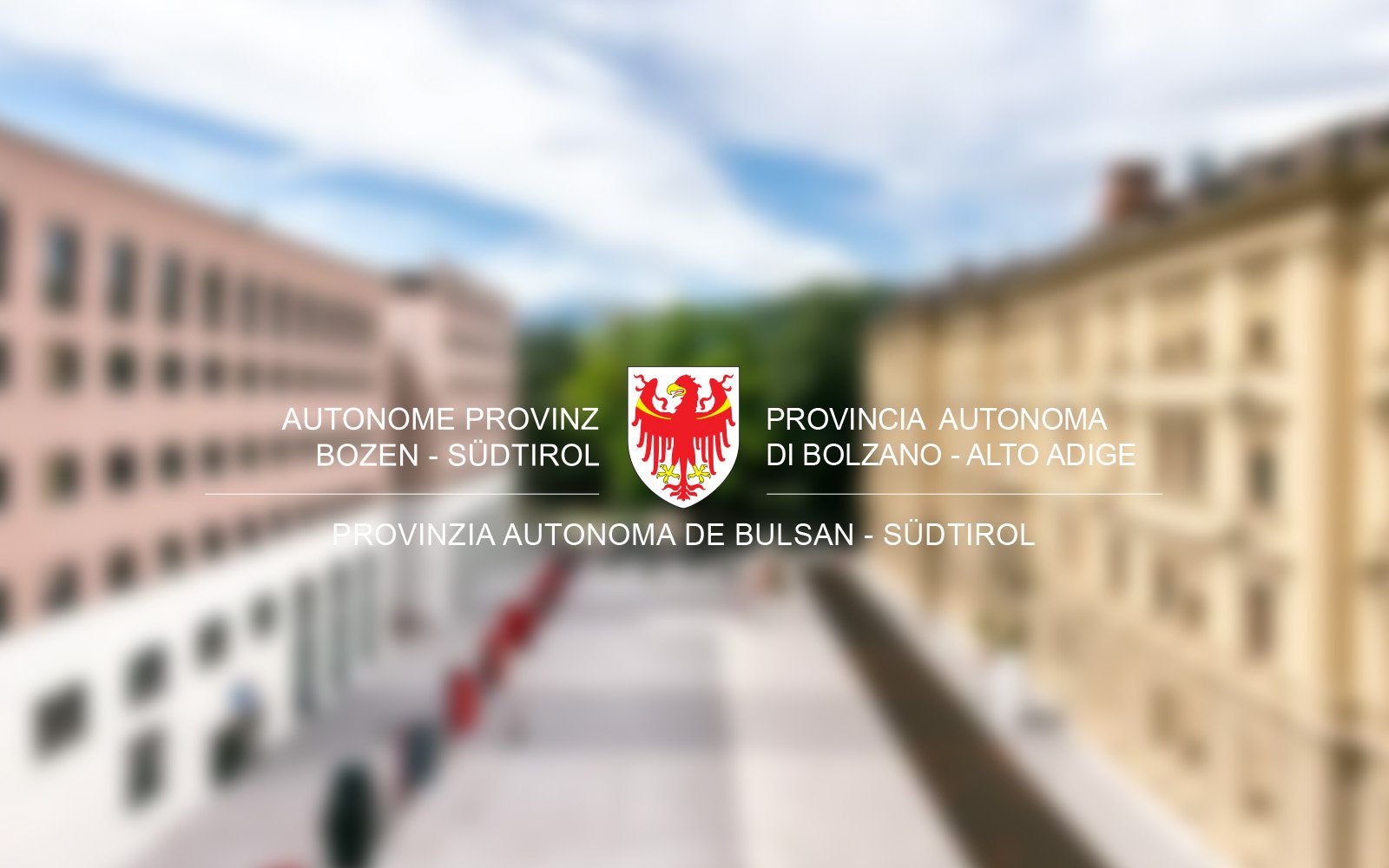 Blurry background of buildings with a logo in the center featuring a red eagle and text in four languages indicating Bolzano, South Tyrol, Italy.